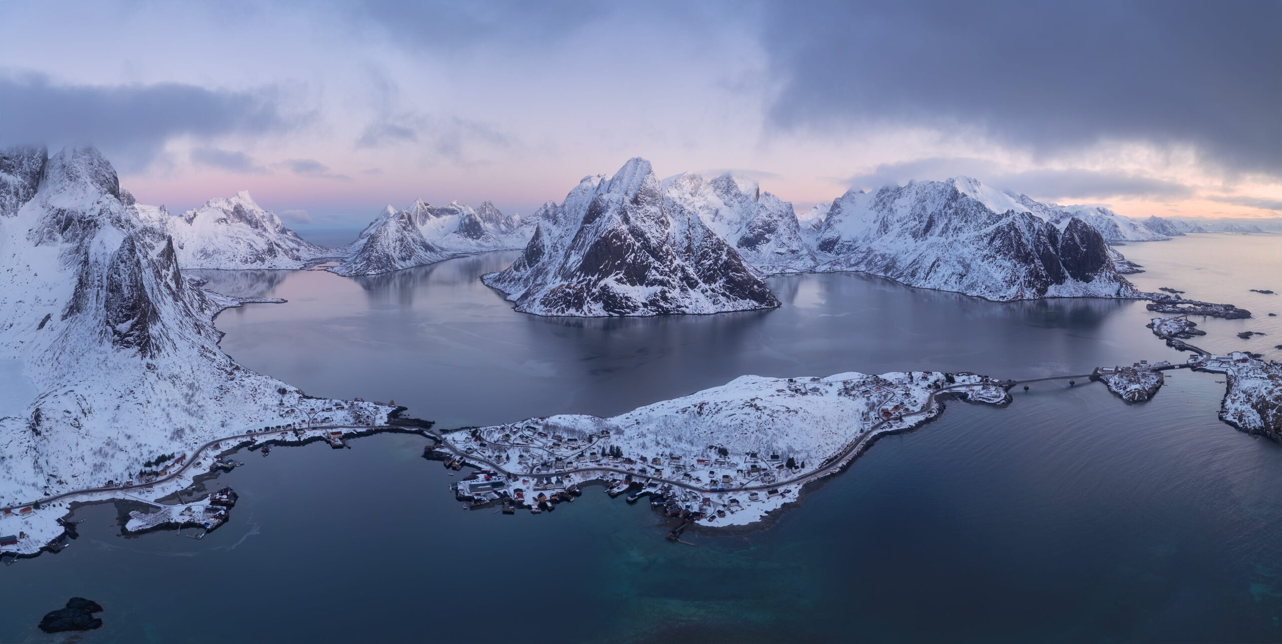 Panoramic aerial landscape photography of Reine at blue hour in winter, Lofoten Islands, Norway, showing the fishing village, fjord and snow-covered mountains under soft Arctic twilight.