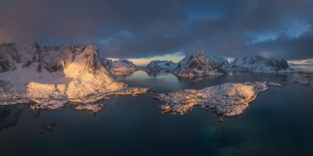 Panoramic aerial drone landscape photography of Reine village at sunrise in winter, Lofoten Islands, Norway, showing snow-covered mountains, fjords and the fishing village illuminated by soft Arctic morning light.