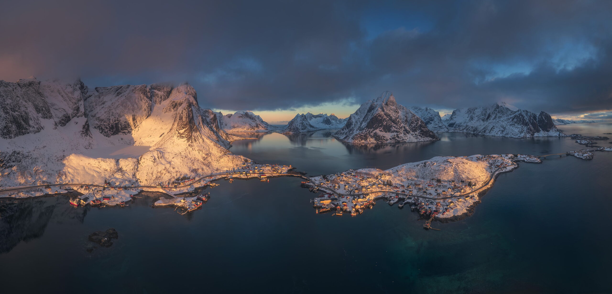 Panoramic aerial drone landscape photography of Reine village at sunrise in winter, Lofoten Islands, Norway, showing snow-covered mountains, fjords and the fishing village illuminated by soft Arctic morning light.