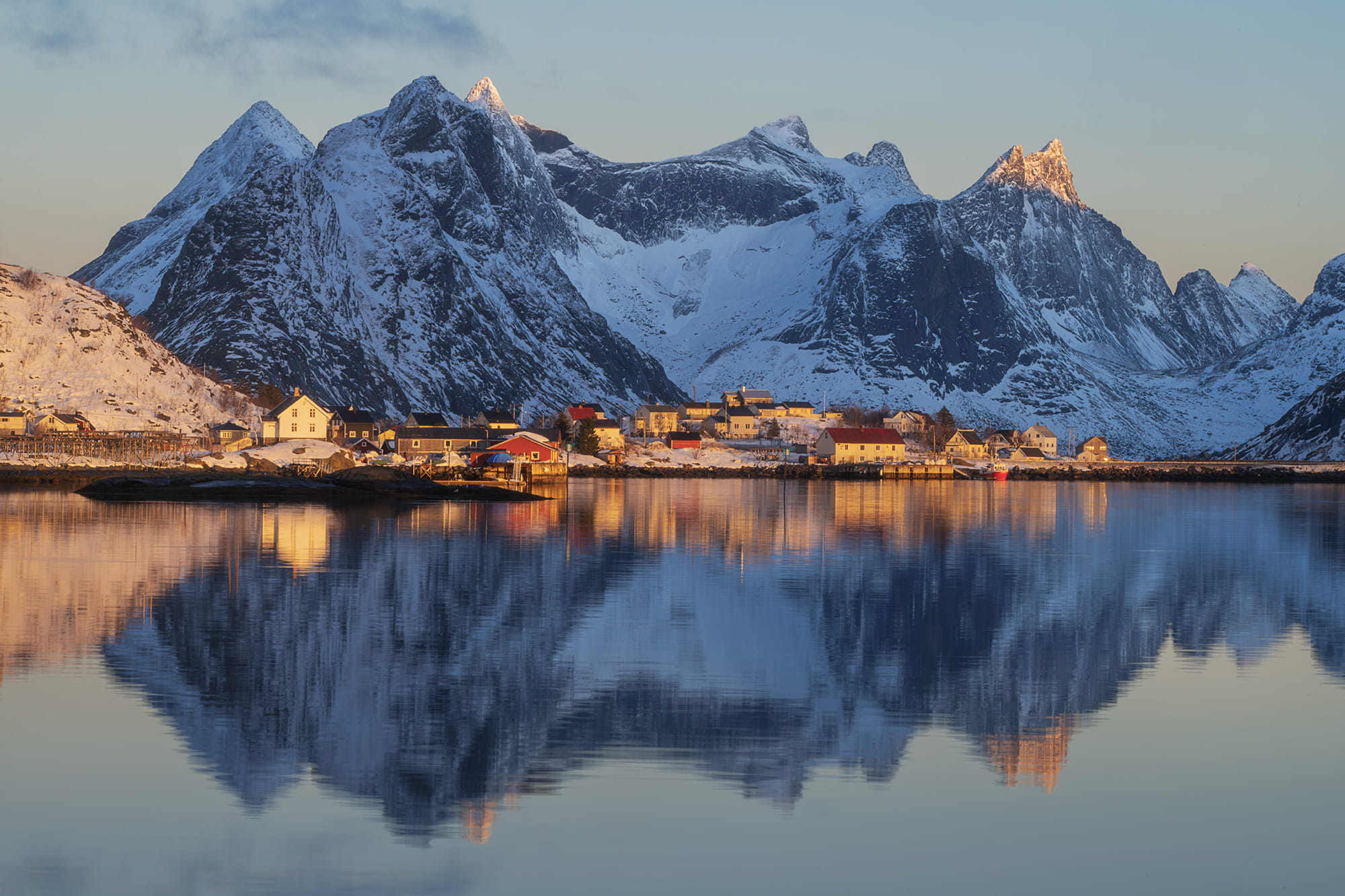 Landscape photography of Reine at sunrise, Lofoten Islands, Norway, with calm fjord reflections and snow-covered mountains glowing in soft Arctic morning light.