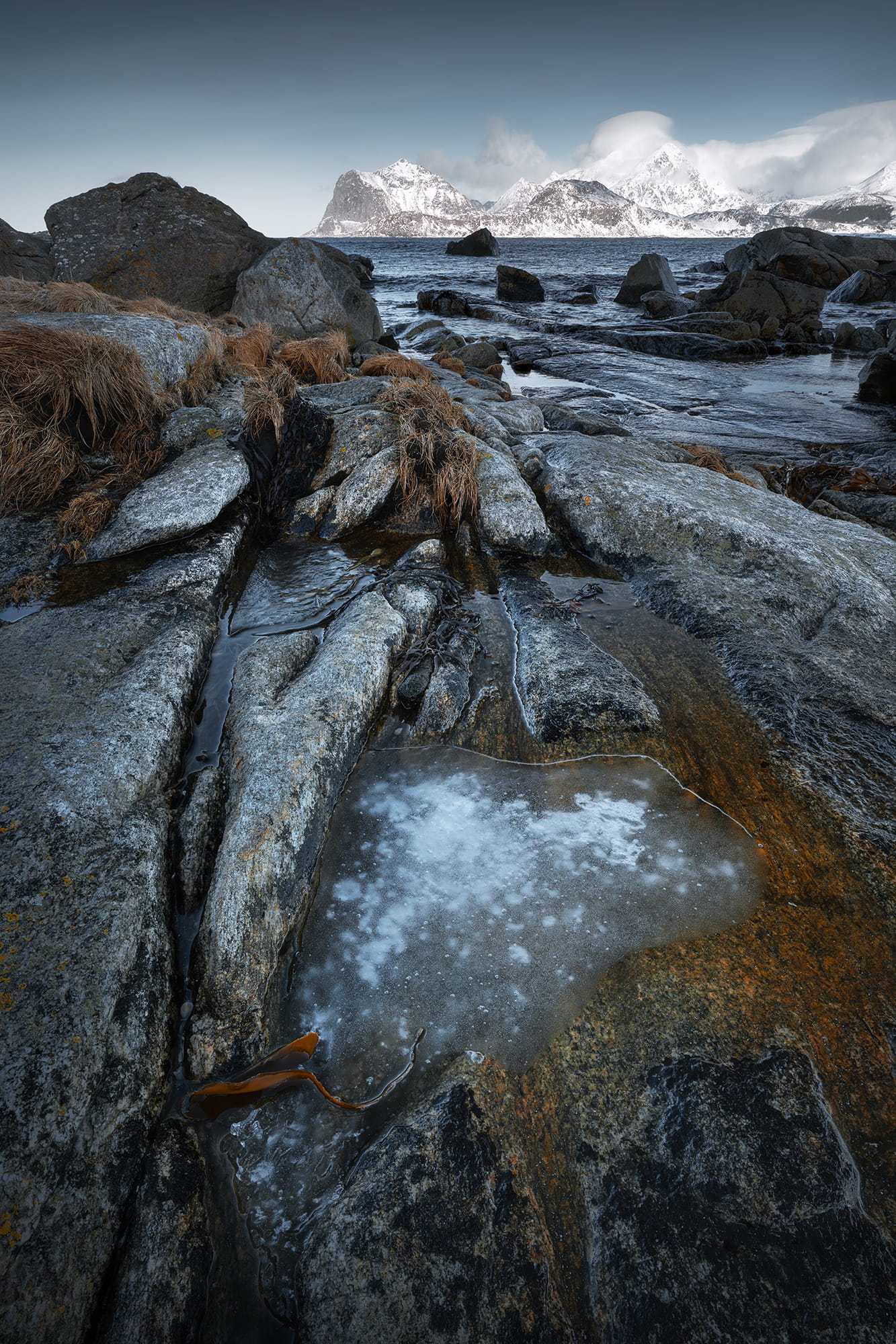 Winter seascape landscape photography near Myrland in the Lofoten Islands, Norway, featuring frozen water, textured rocks and snow-covered mountains along the Arctic coast.