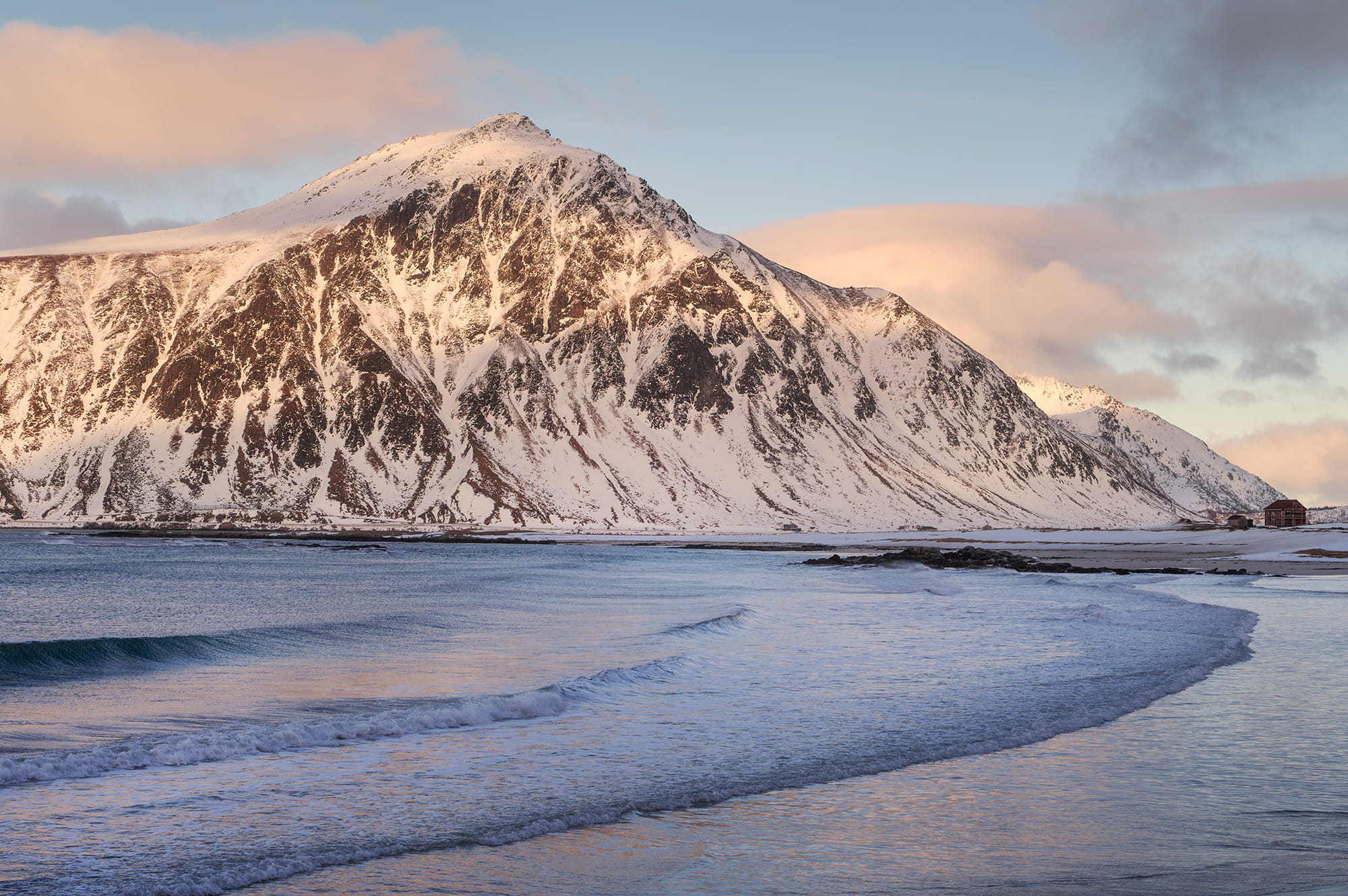 Winter landscape photography of Skagsanden Beach in Flakstad, Lofoten Islands, Norway, with snow-covered mountain, soft waves and pastel light at sunset.