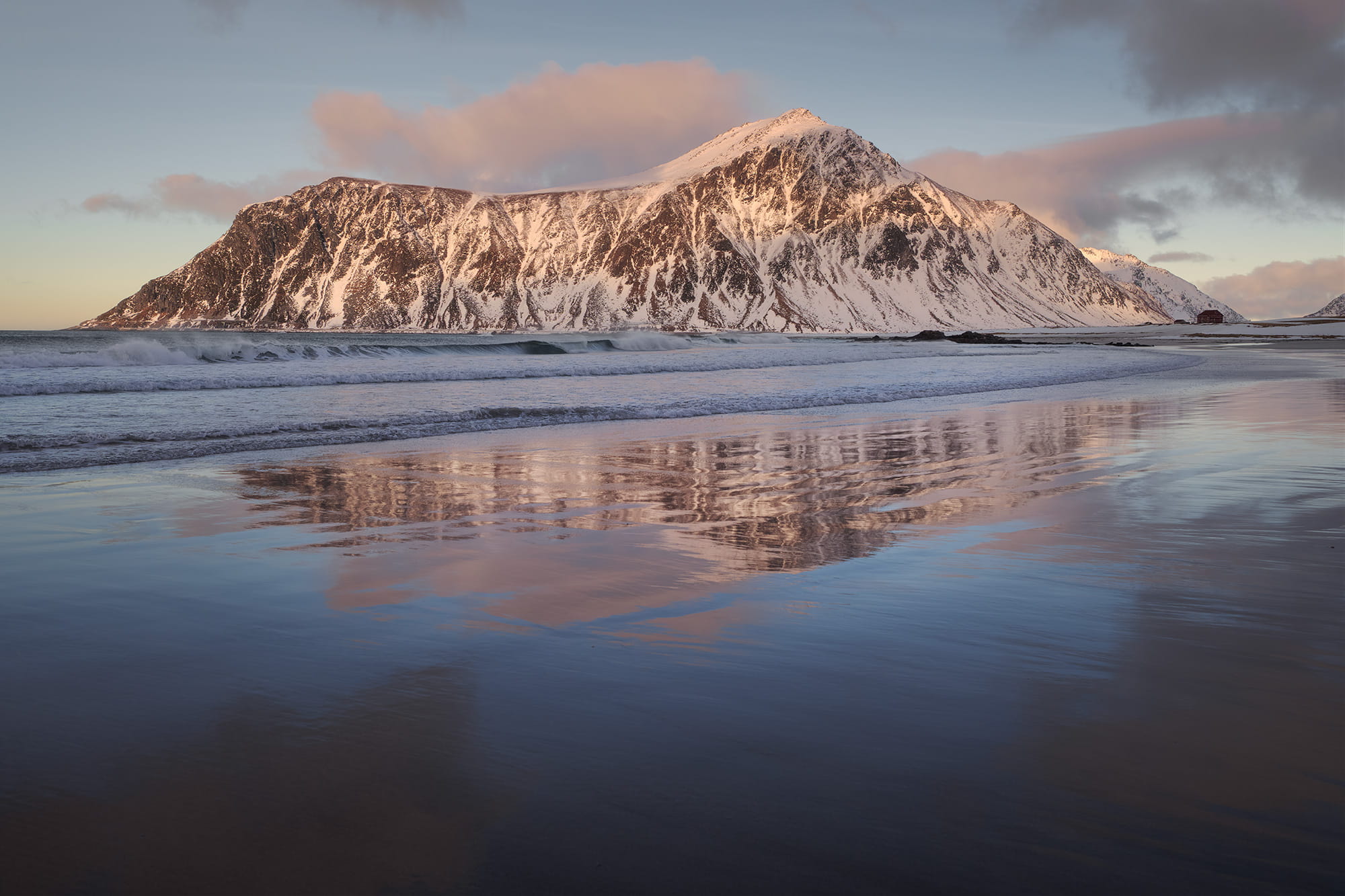 Sunset reflection on Skagsanden Beach in Flakstad, Lofoten Islands, Norway, with snow-covered mountains and waves reflecting on wet sand during winter.
