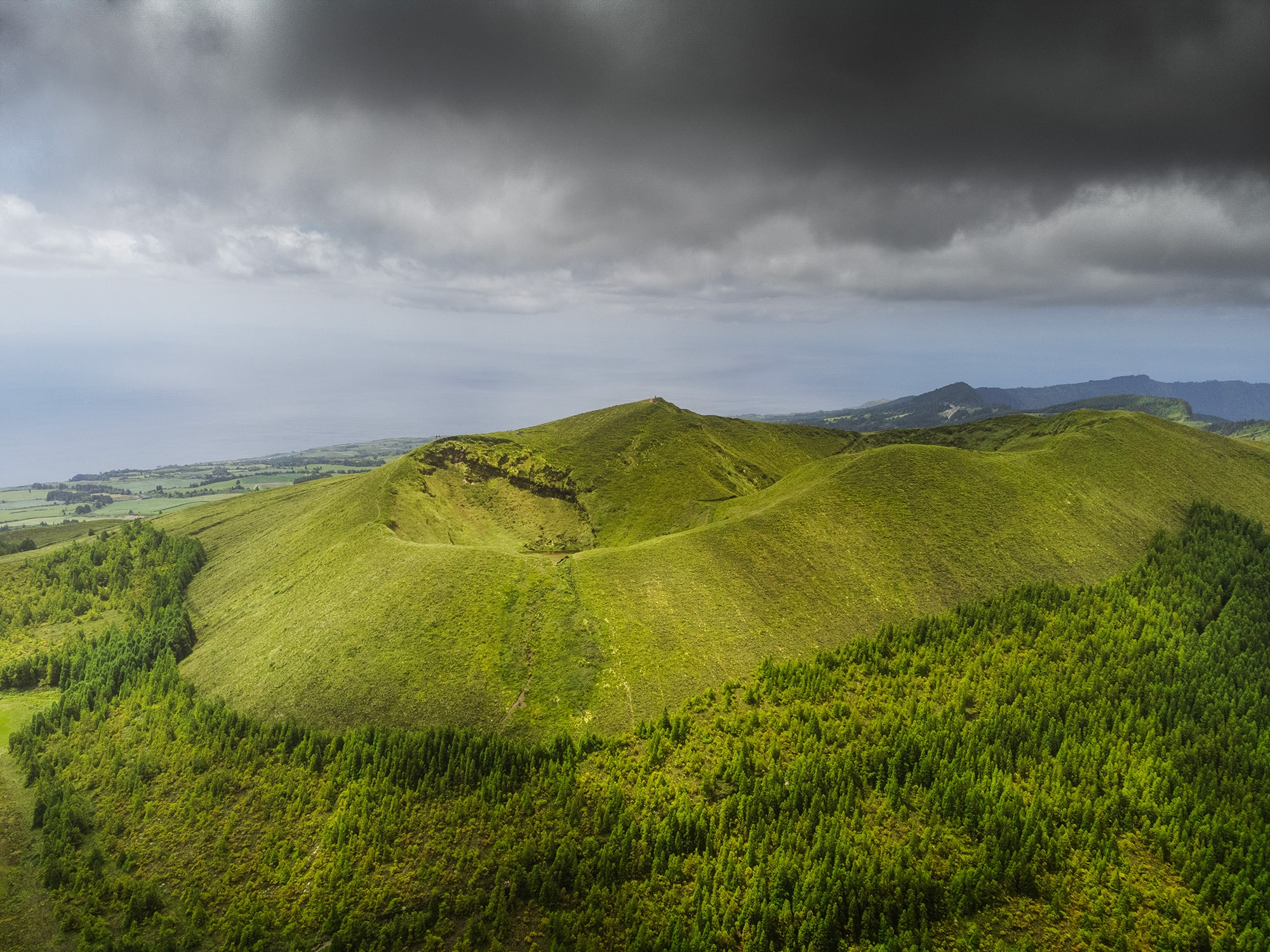 Aerial view of Pico das Éguas volcanic crater on São Miguel Island, Azores, with lush green hills, dense forest and dramatic cloudy sky over the Atlantic coastline.