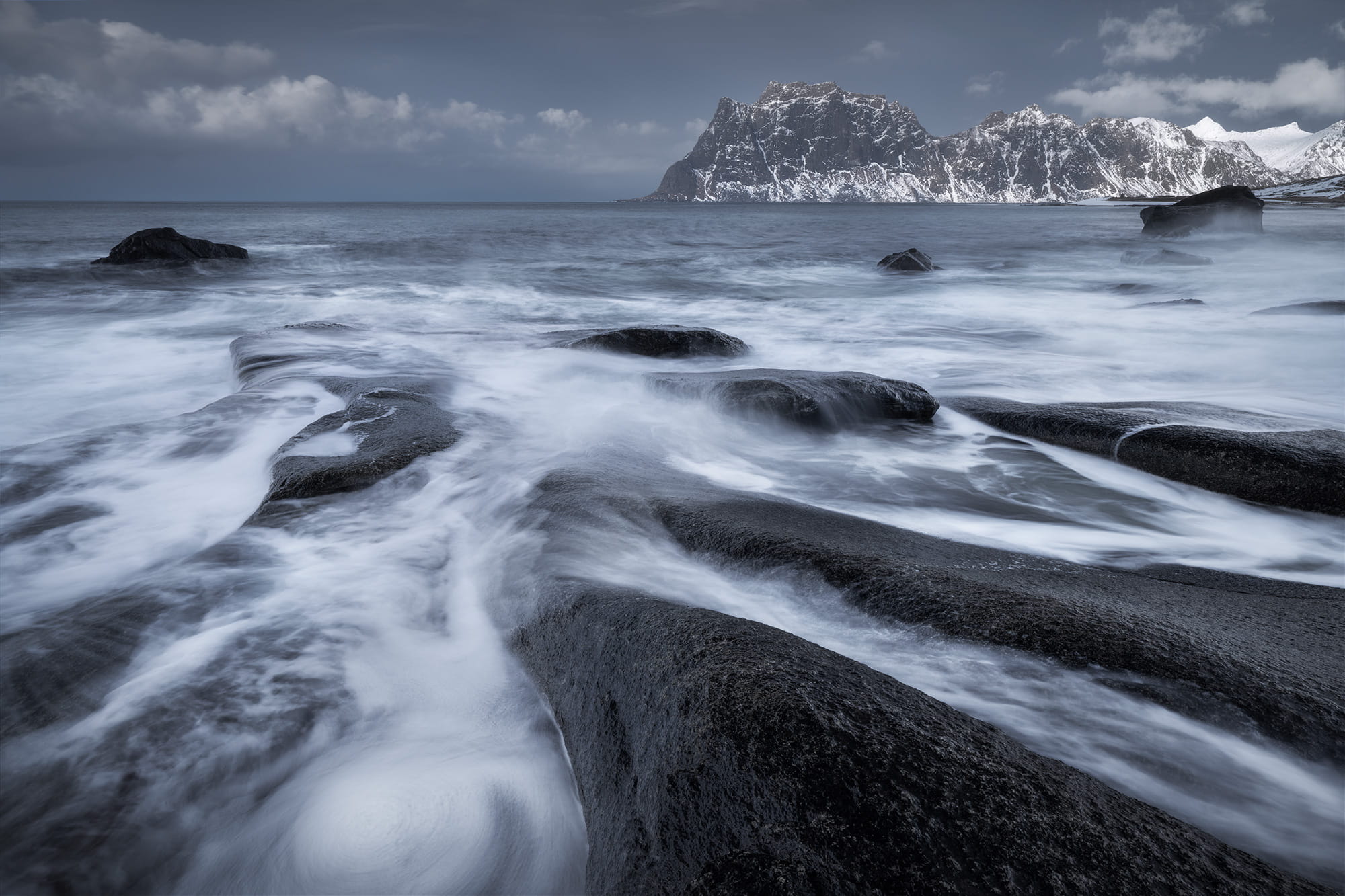 Long exposure seascape at Uttakleiv Beach in the Lofoten Islands, Norway, with smooth flowing water over dark rocks leading toward snowy mountains in a winter landscape.