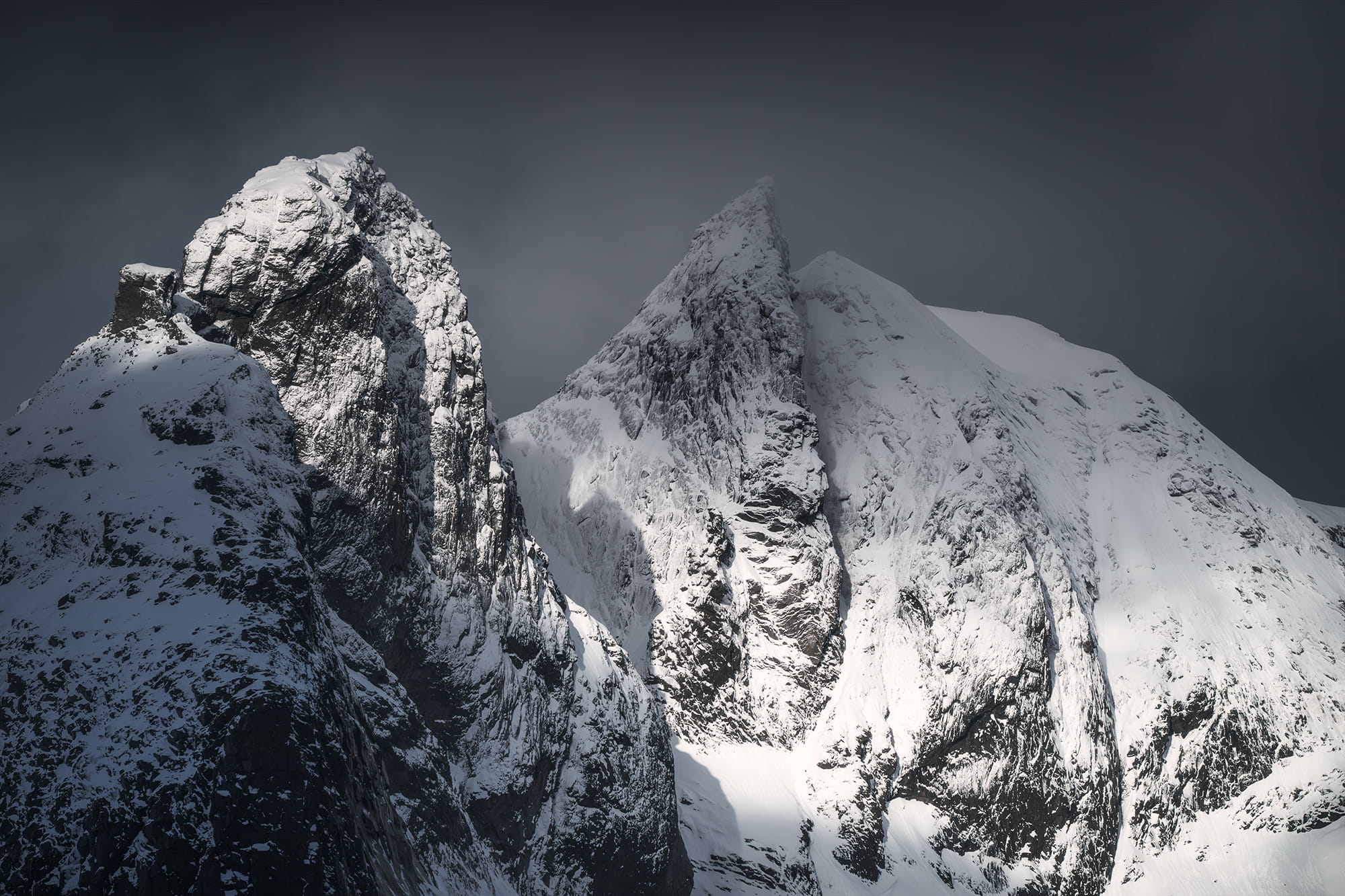 Snow-covered peaks near Reine in the Lofoten Islands, Norway, captured in dramatic light with deep shadows highlighting the rugged mountain textures in winter.