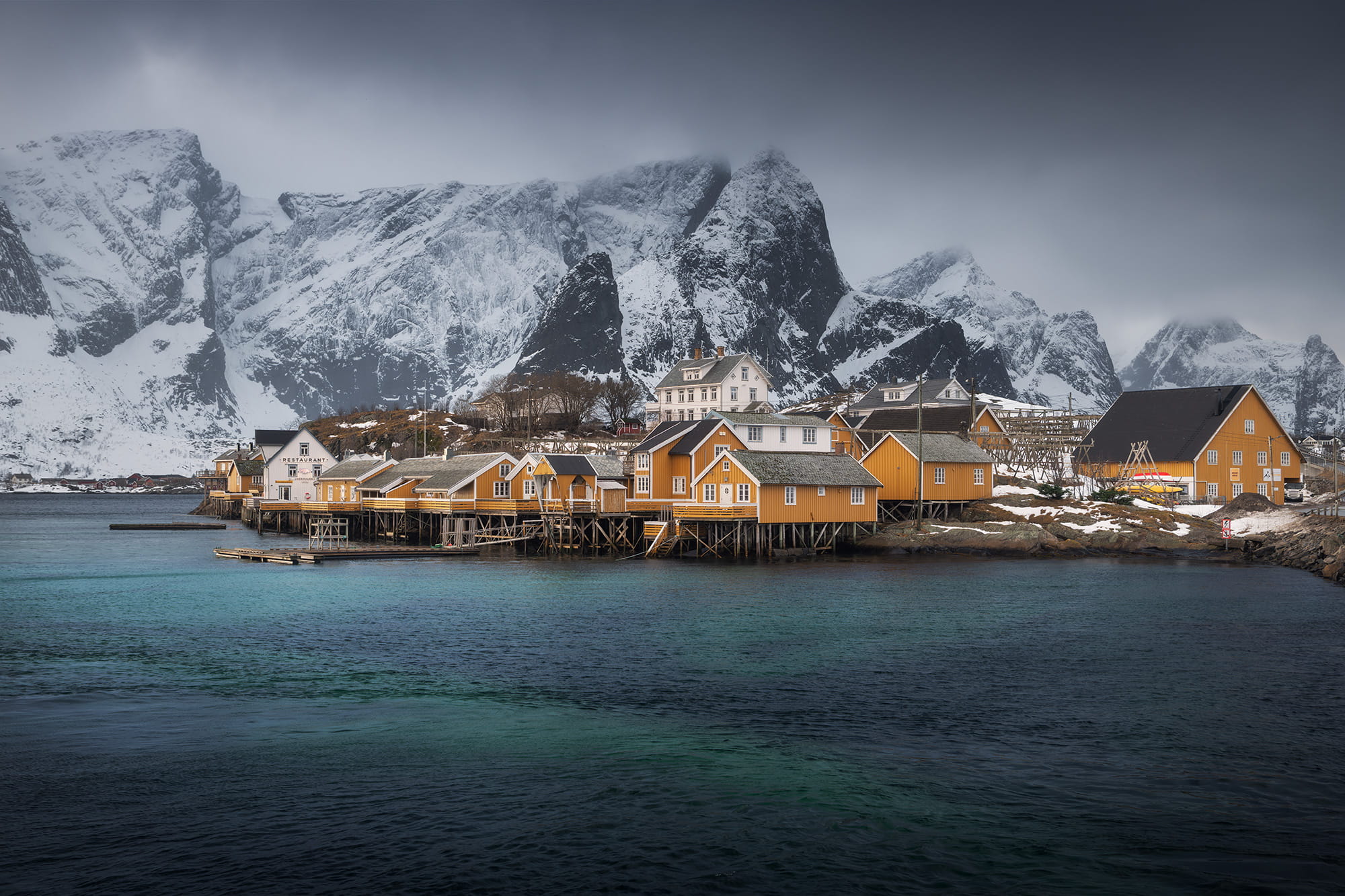 Hamnøy fishing village in winter in the Lofoten Islands, Norway, with traditional yellow rorbuer cabins on stilts and snow-covered mountains in the background.