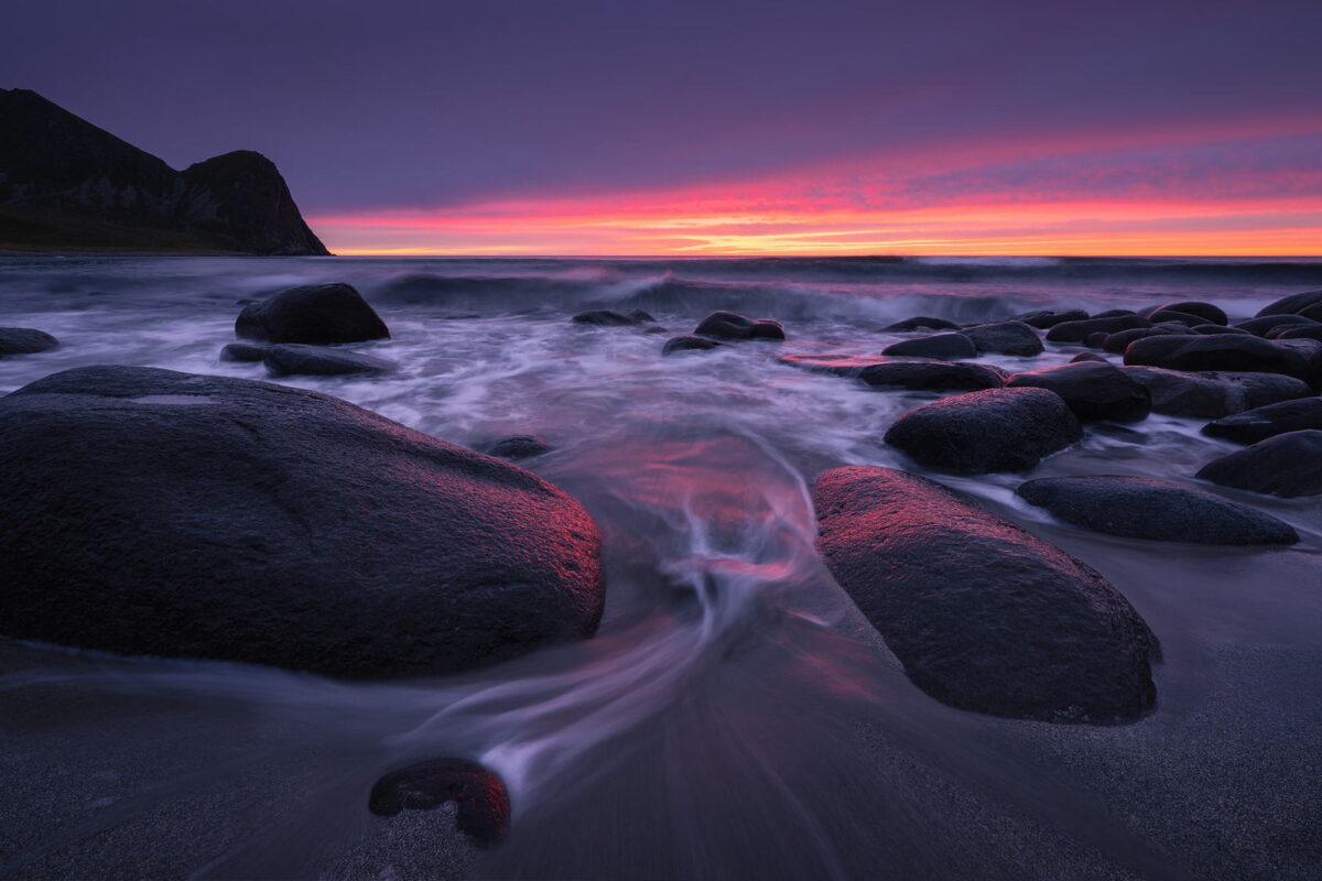 Long-exposure landscape photography of a red sunset at Unstad Beach in the Lofoten, Norway, featuring a heart-shaped rock in the foreground under dramatic sunset.