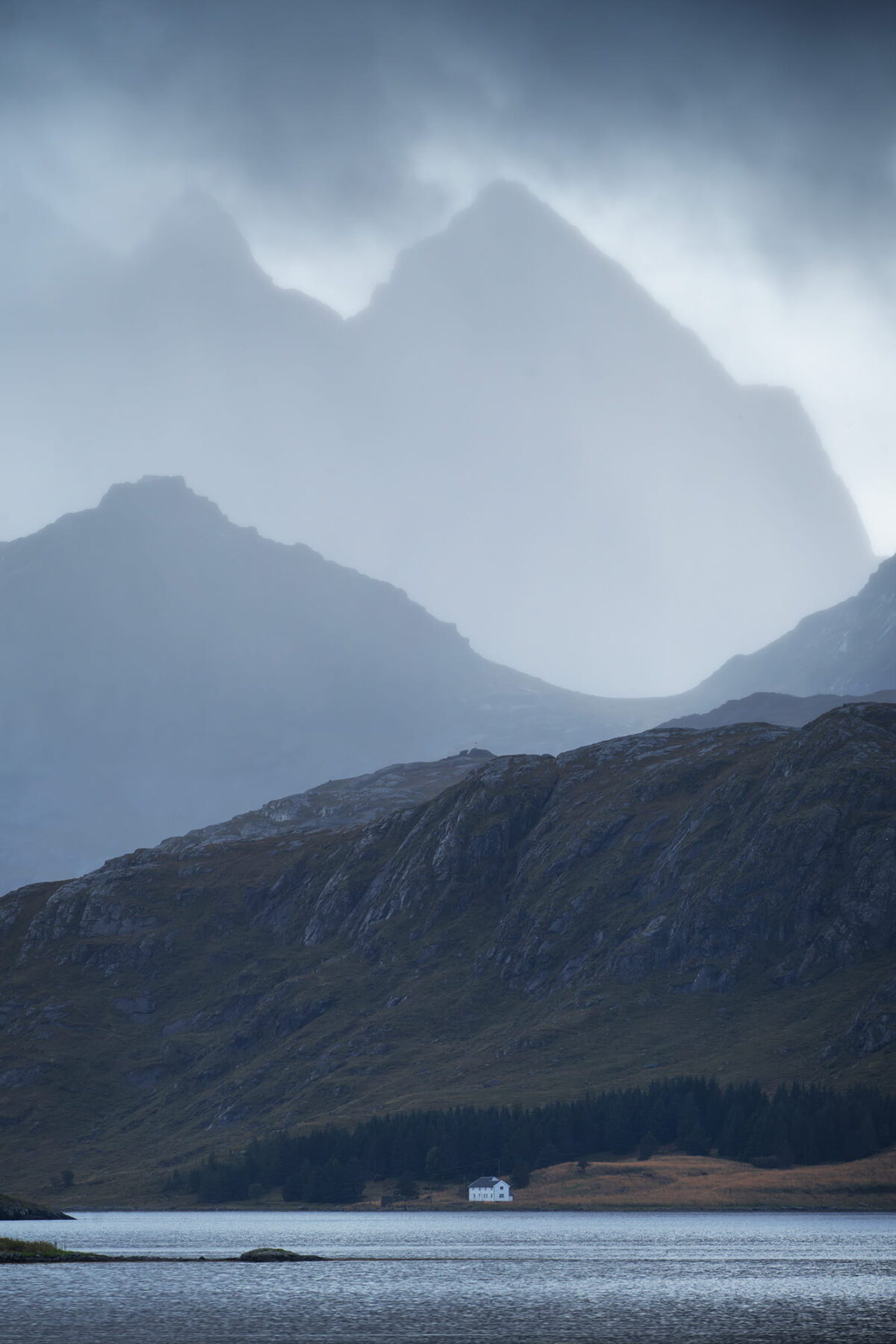 Moody landscape at Fredvang in the Lofoten Islands, Norway, with foggy mountains, fjord and a small white house under dramatic sky.