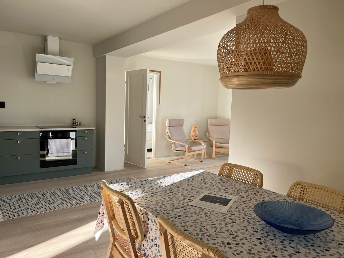 Bright kitchen and dining area in an apartment in central Lofoten near Gravdal, with a wooden table, woven chairs, a modern green kitchen and soft natural light coming from the side.