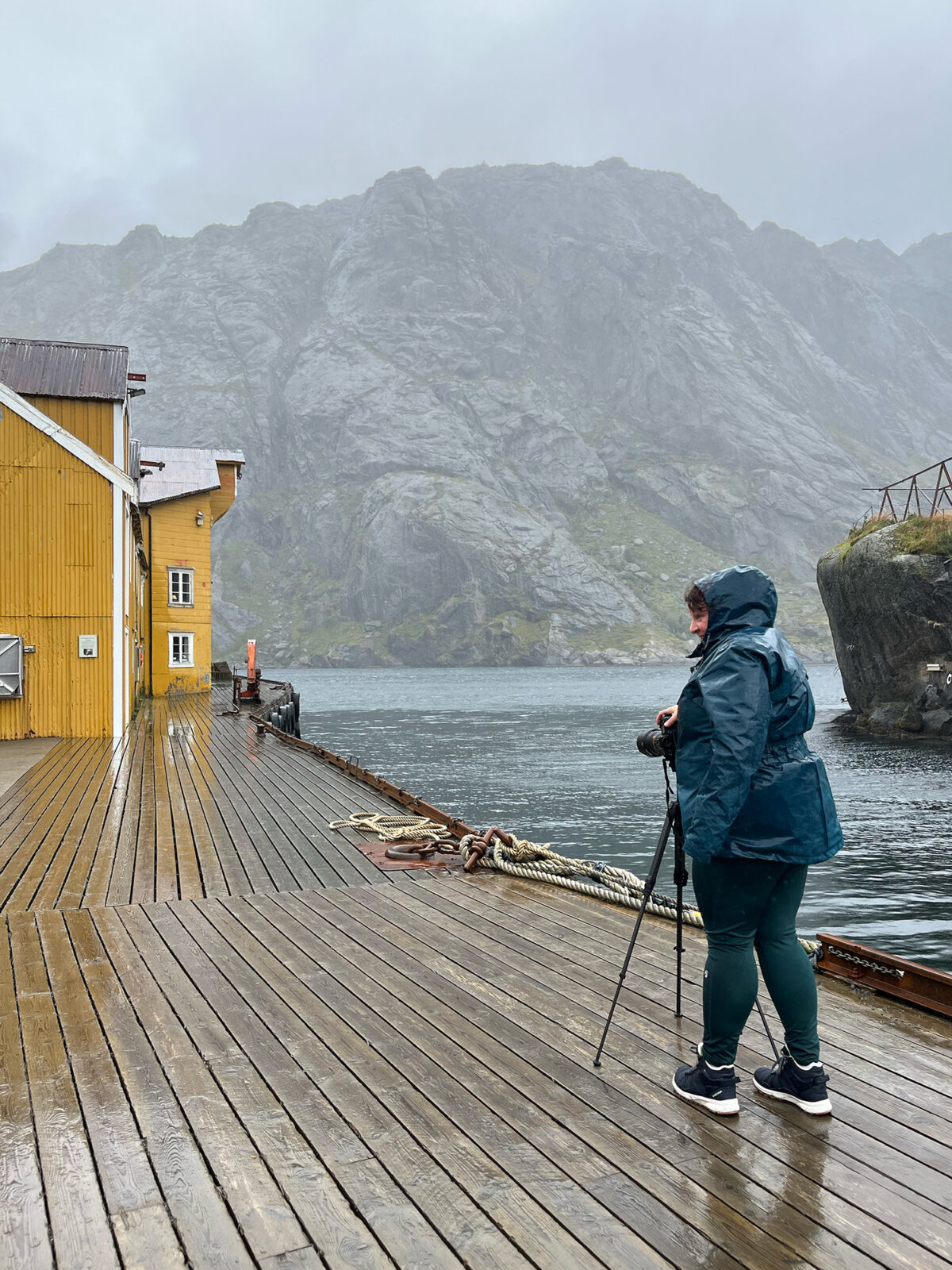 Landscape photographer photographing a yellow fisherman’s house in Nusfjord in the rain, Lofoten Islands, Norway