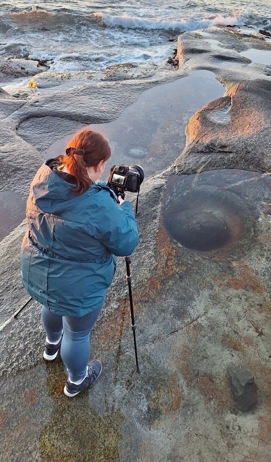 Jennifer Esseiva photographing the Dragon Eye rock at Uttakleiv Beach in the Lofoten Islands, Norway, wearing a turquoise jacket.
