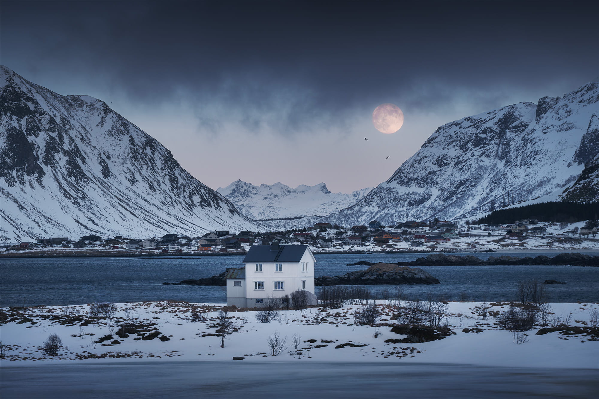 Full moon rising above snowy mountains and the village of Ramberg in the Lofoten Islands, Norway, captured during blue hour with a 70-200mm telephoto lens. Arctic winter landscape with fjord, dramatic peaks and a white house in the foreground.