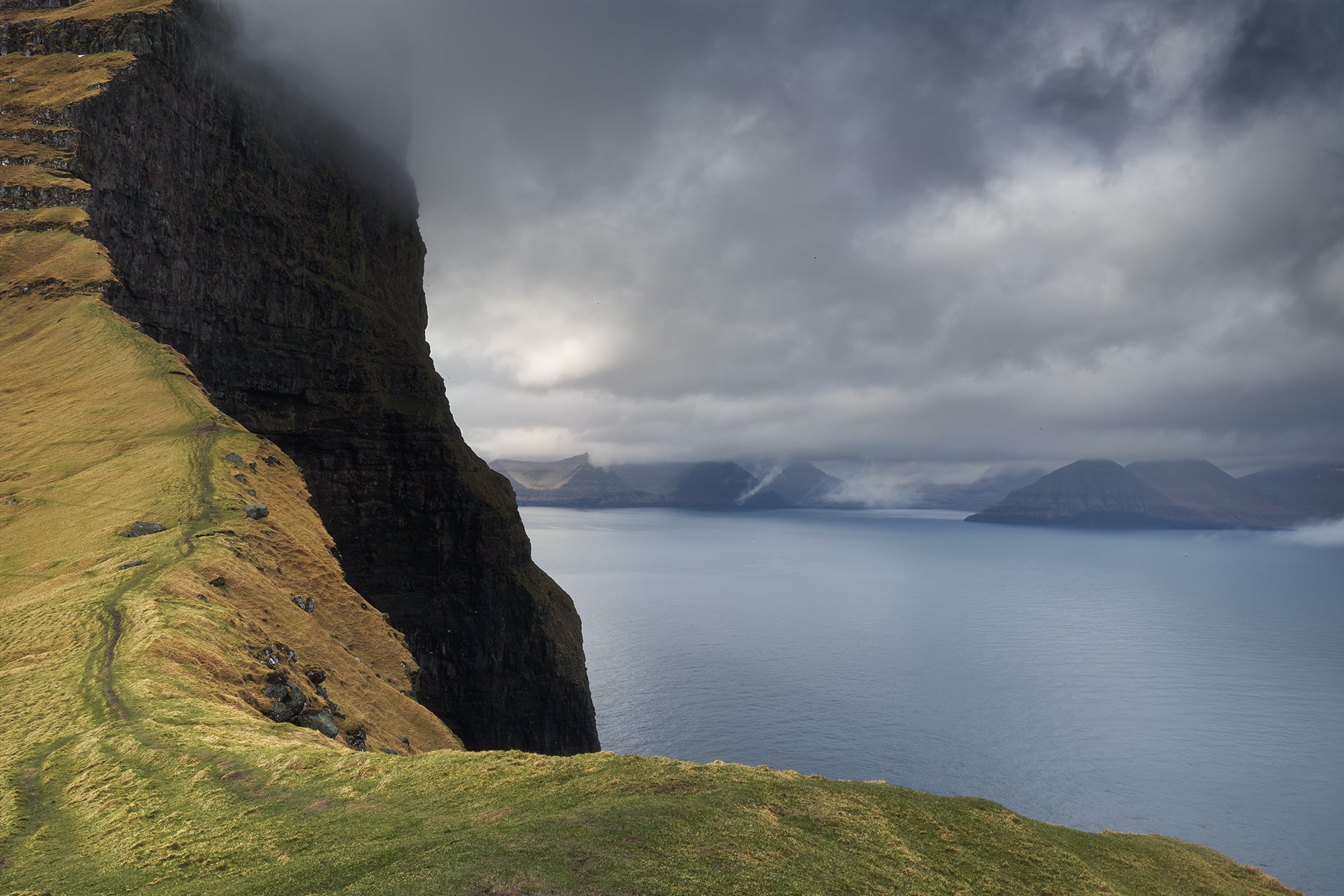 Dramatic coastal cliffs on Kalsoy Island in the Faroe Islands, with a narrow hiking trail overlooking the North Atlantic beneath moody storm clouds.