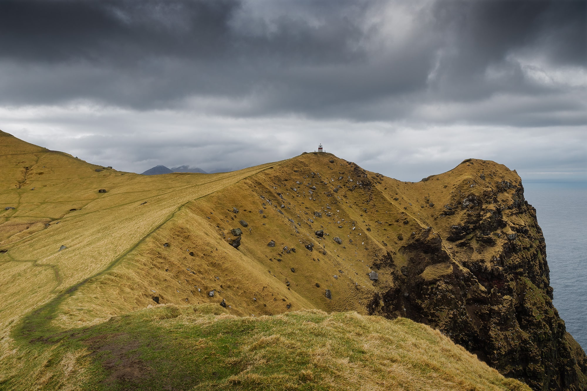 Kallur Lighthouse on Kalsoy Island in the Faroe Islands, surrounded by dramatic coastal cliffs and rolling green ridges beneath a moody overcast sky.