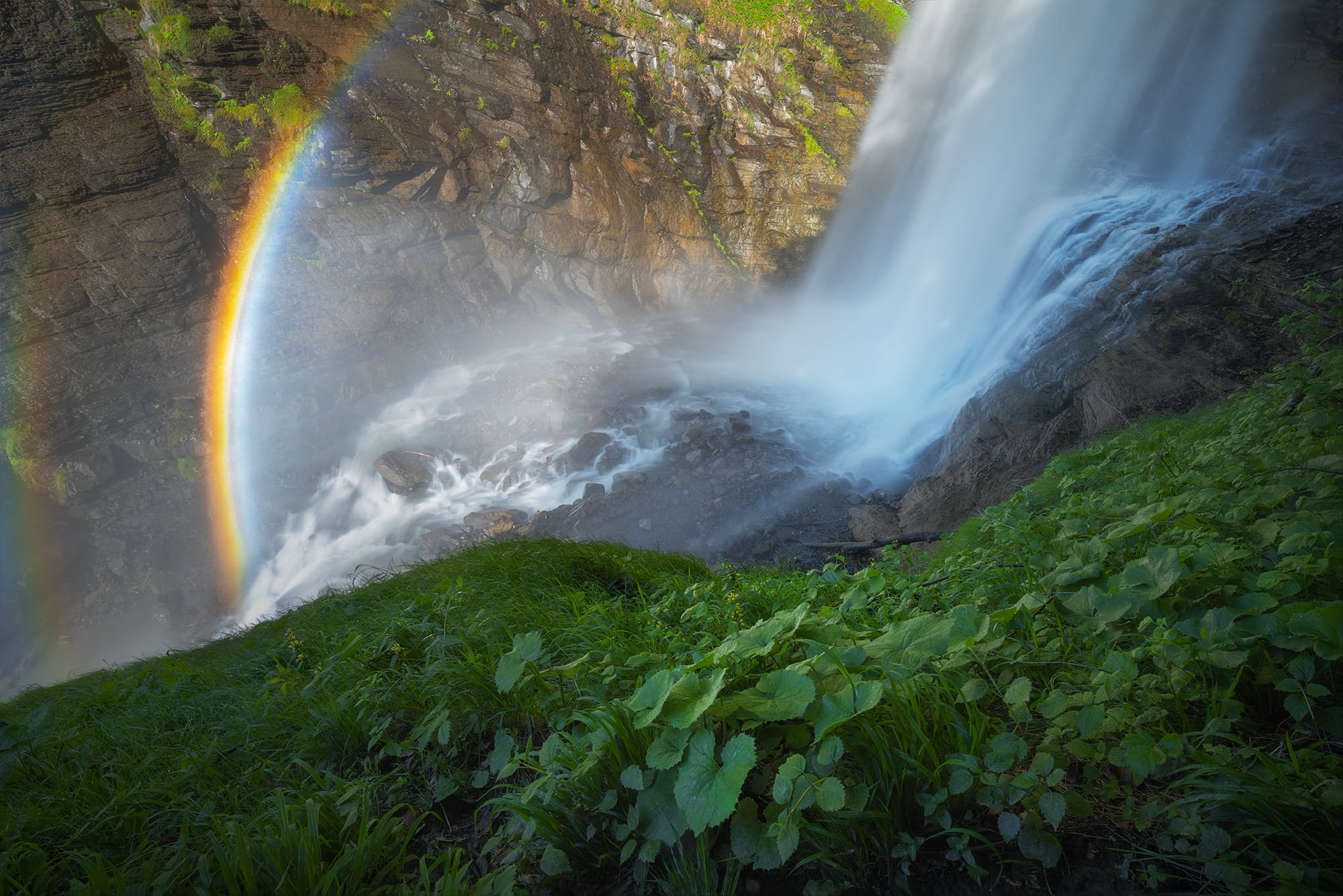 Rainbow at Diesbachfall waterfall in the Canton of Glarus, Switzerland. Long exposure photograph showing the powerful waterfall, lush green vegetation and a bright rainbow forming in the mist on a sunny afternoon.