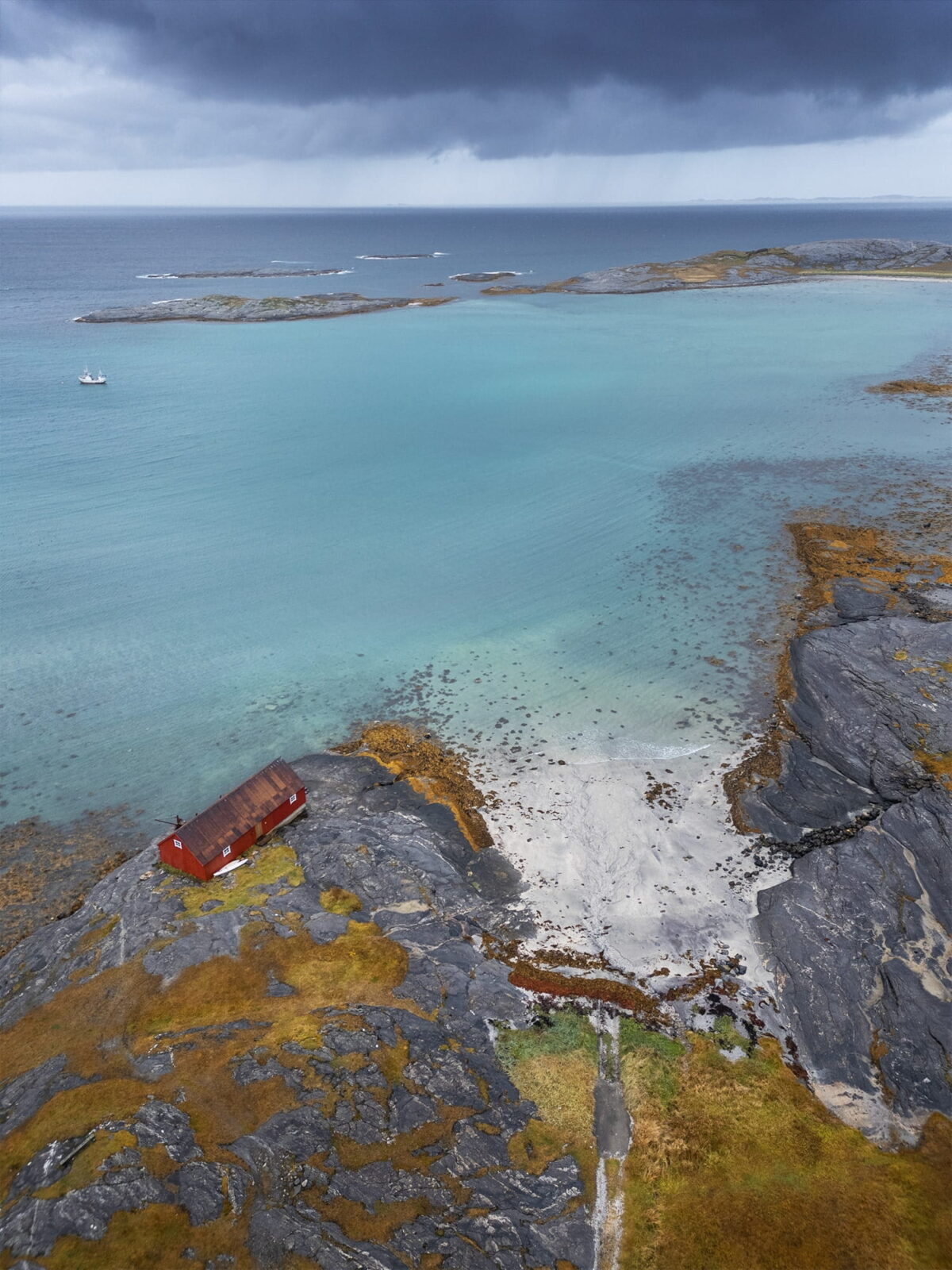 Fine art aerial photograph of a red fishing cabin on the rocky coastline near Mjelle Beach, Bodø, Northern Norway, with turquoise Arctic waters and approaching storm clouds.