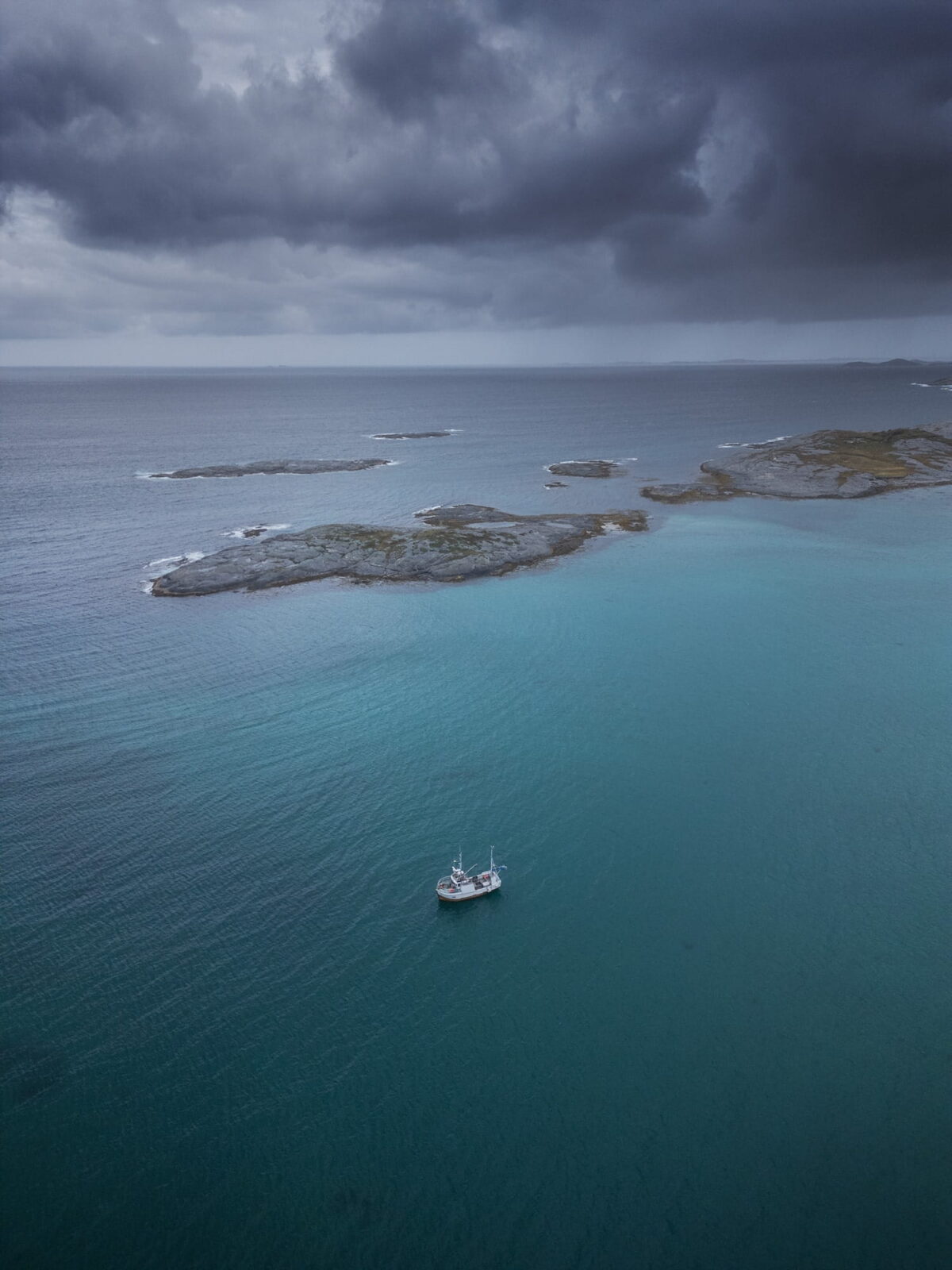 Drone photograph of a fishing boat floating on turquoise Arctic waters near Mjelle Beach, Bodø, Northern Norway, beneath dramatic storm clouds and rocky islands.