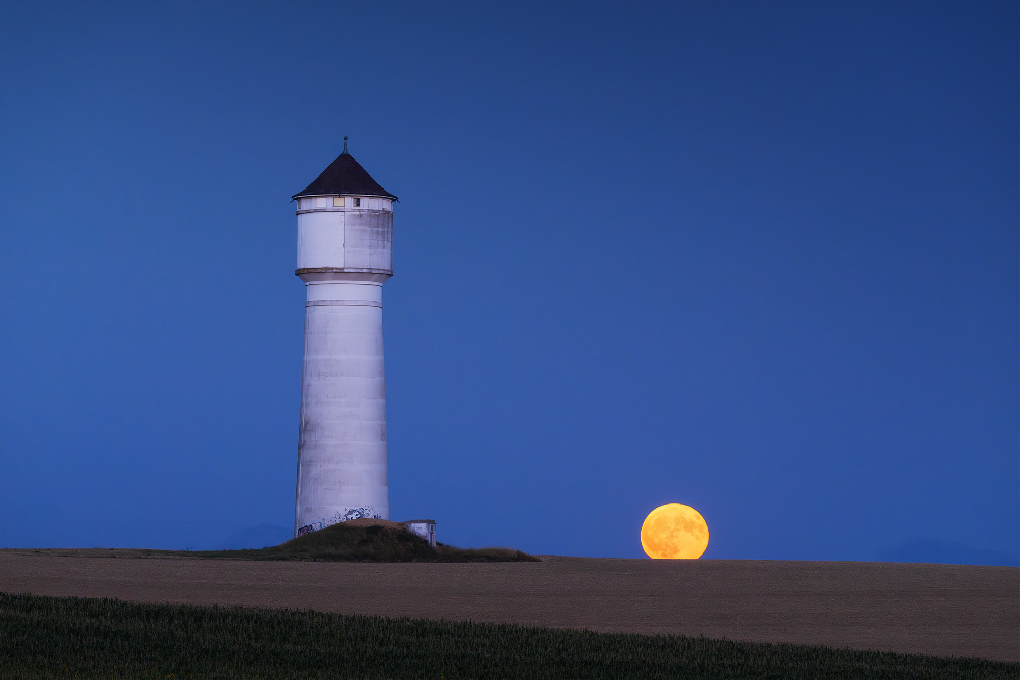 Fine art landscape photograph of the Echallens water tower in the Canton of Vaud, Switzerland, with a full moon rising at dusk beneath a deep blue twilight sky.