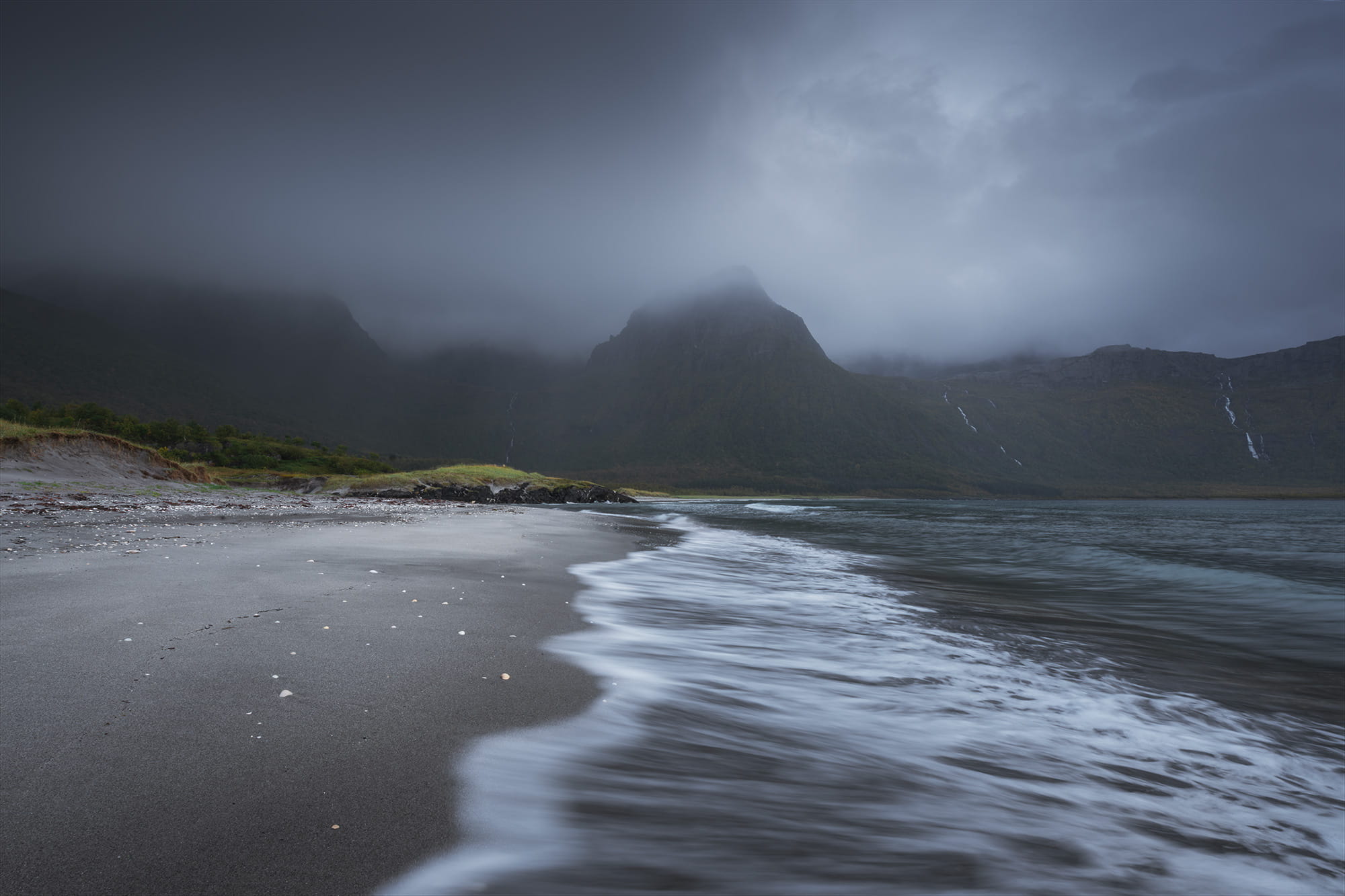 Moody seascape at Brennviksanden Beach in Steigen, Northern Norway, with gentle waves washing over a black sand beach beneath mist-covered mountains and dramatic storm clouds.