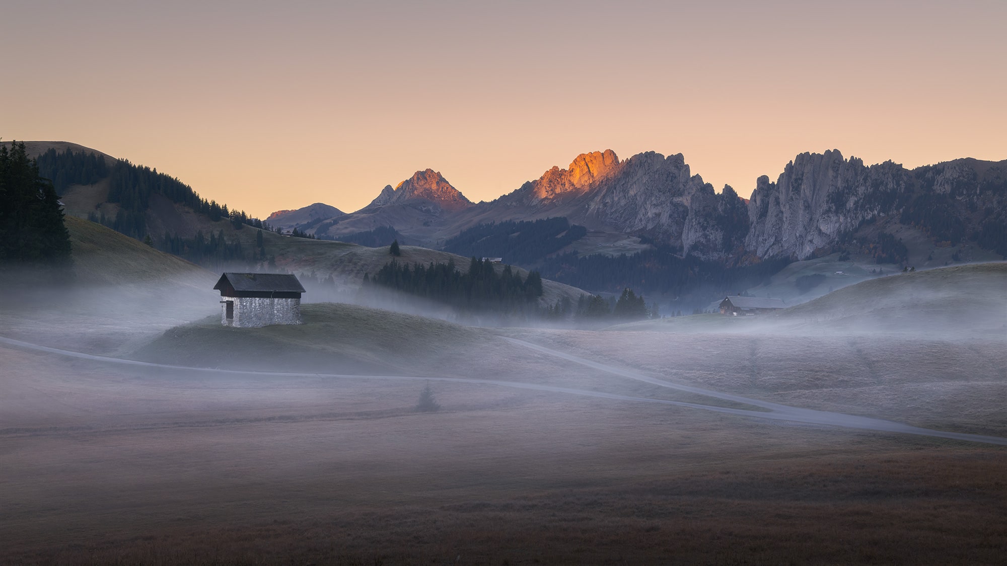 Autumn sunrise over the Gastlosen mountain range from Jaun Pass, Switzerland, with golden alpine peaks emerging above morning mist and a solitary mountain hut.
