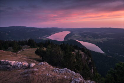 Panoramic sunset over Lac de Joux and Lac des Brenets from Dent de Vaulion, Switzerland, with vibrant pink skies reflected on the lakes and layered Jura mountains.