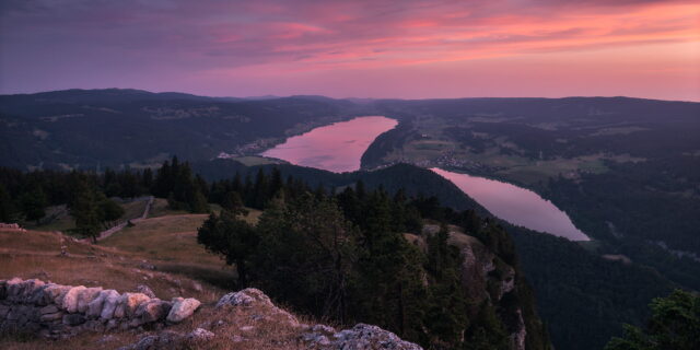 Panoramic sunset over Lac de Joux and Lac des Brenets from Dent de Vaulion, Switzerland, with vibrant pink skies reflected on the lakes and layered Jura mountains.