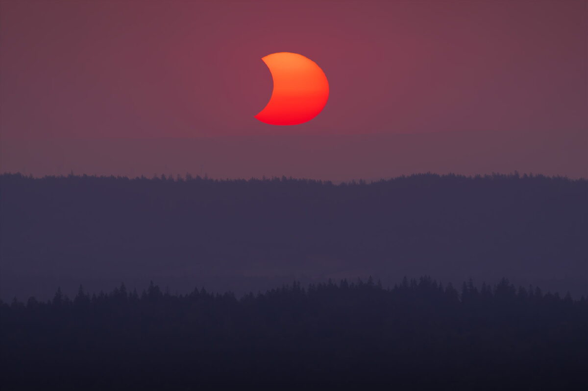 Partial solar eclipse at sunset on August 12, 2026, photographed from Dent de Vaulion above the Vallée de Joux in the Swiss Jura, Switzerland, with the eclipsed red Sun glowing above layered forested hills.