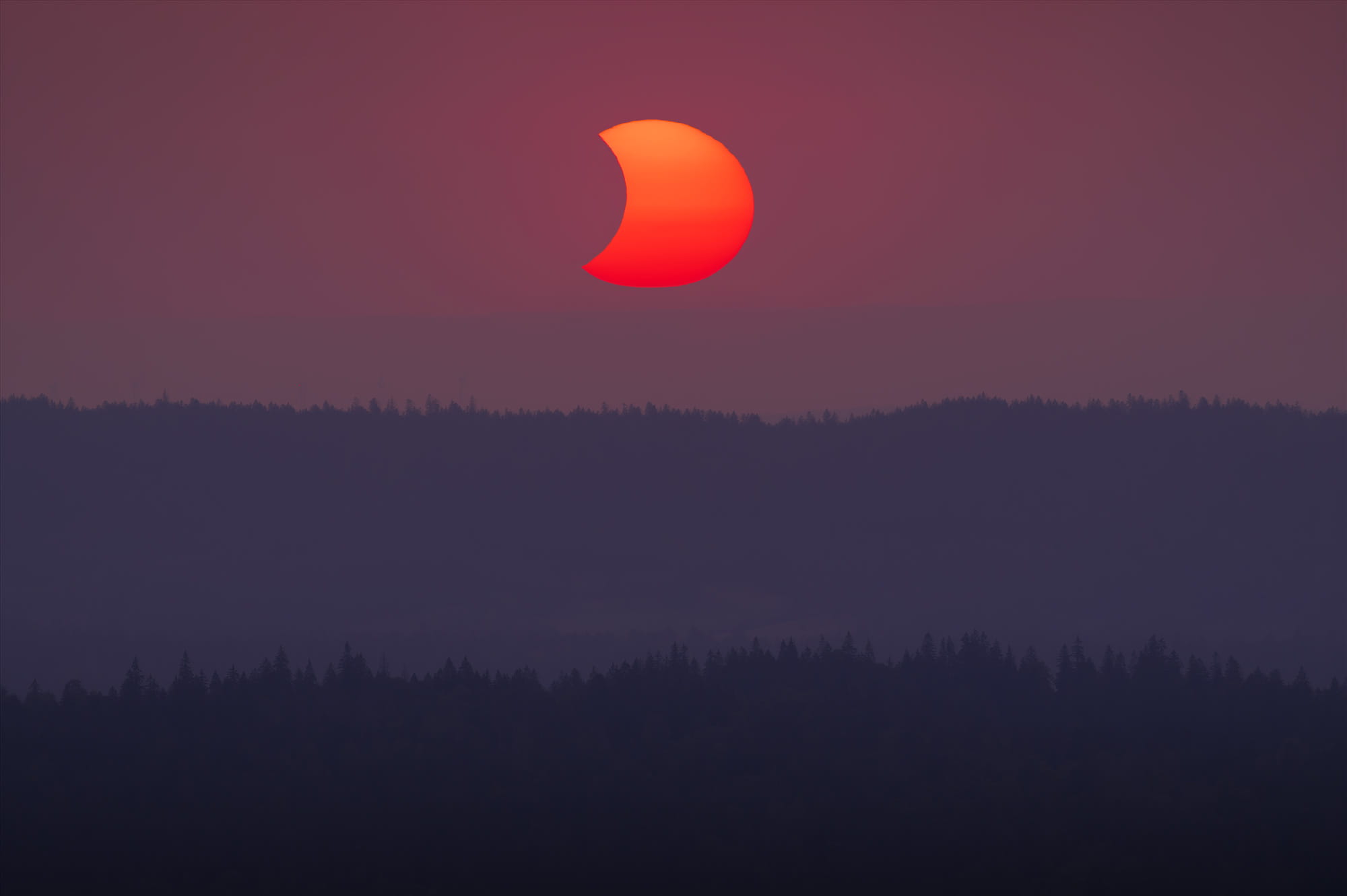 Partial solar eclipse at sunset on August 12, 2026, photographed from Dent de Vaulion above the Vallée de Joux in the Swiss Jura, Switzerland, with the eclipsed red Sun glowing above layered forested hills.