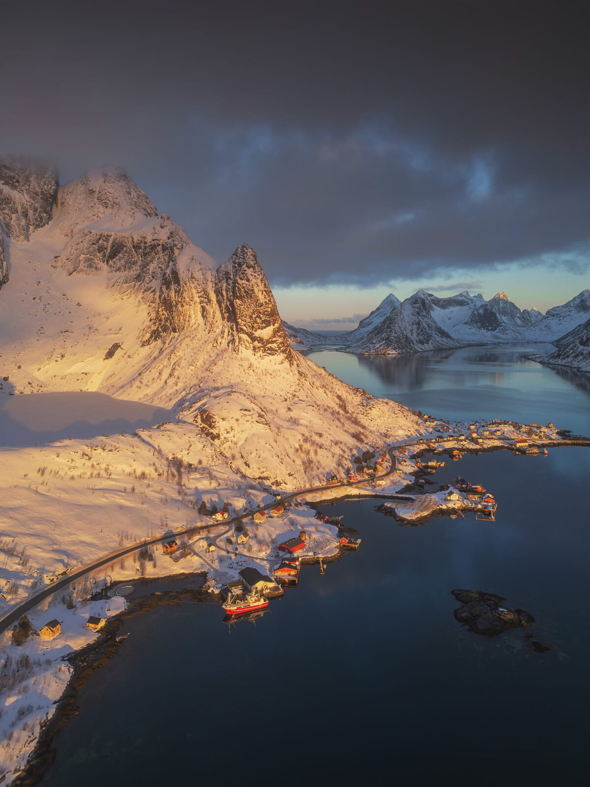 Aerial winter view of Reine in the Lofoten Islands, Norway, surrounded by snow-covered mountains and fjords illuminated by golden light.