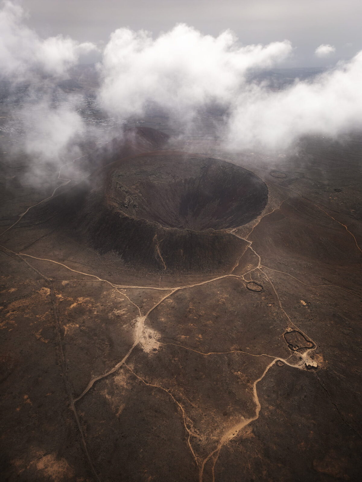 Aerial view of Calderón Hondo volcano in Fuerteventura, with its impressive volcanic crater surrounded by drifting clouds and the arid landscape of the Canary Islands.