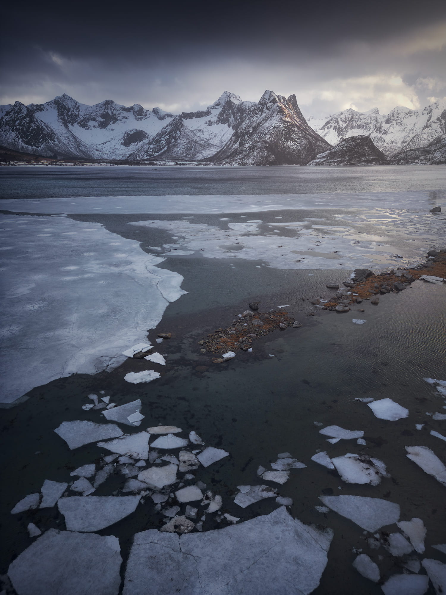 Drone view of Grunnførfjord in winter, with broken sea ice and snow-covered mountains beneath a dramatic sky in the Lofoten Islands, Norway.
