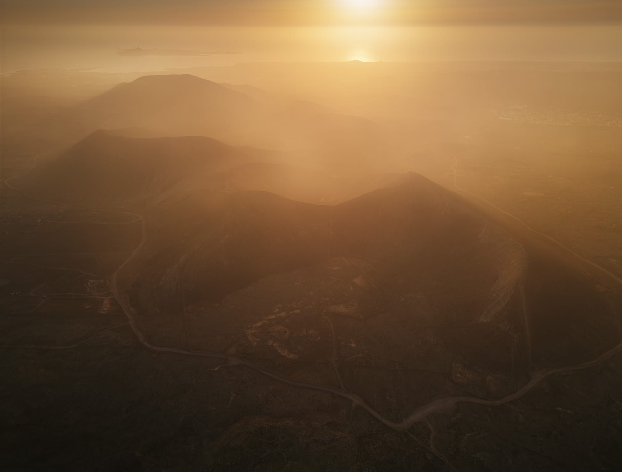 Aerial view of the Bayuyo volcanic chain in Fuerteventura at golden hour, with ancient volcanic craters fading into the warm Atlantic haze near Corralejo, Canary Islands.