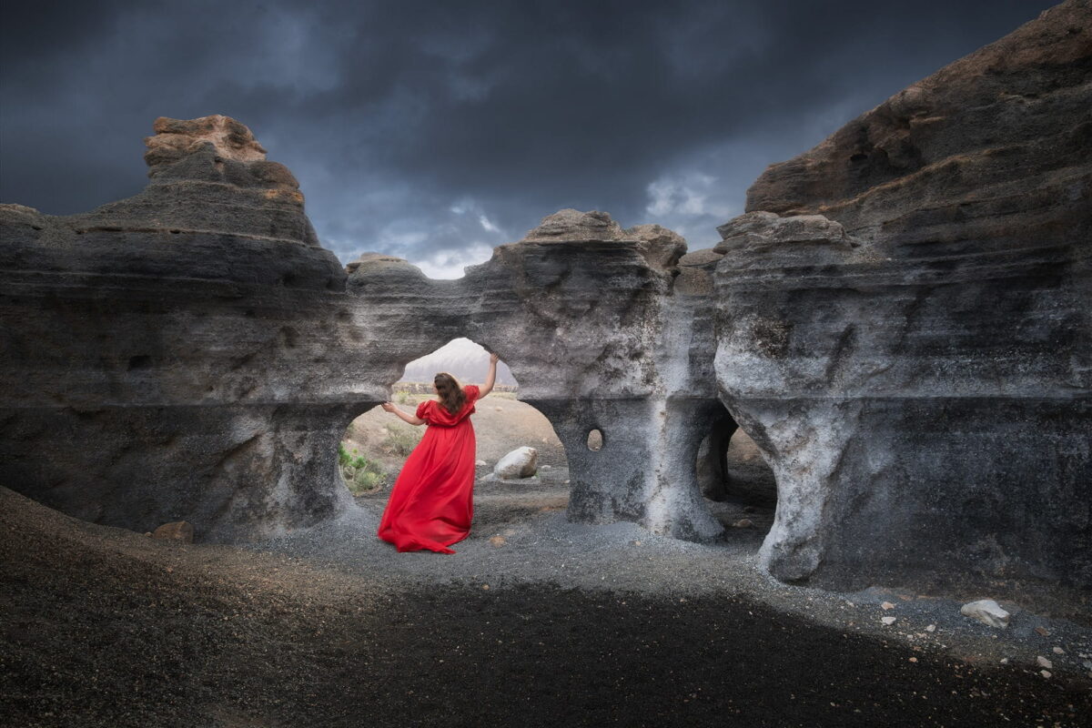 Woman in a flowing red dress among the eroded volcanic rock formations of Ciudad Estratificada in Lanzarote, Canary Islands, Spain, beneath a dramatic cloudy sky.