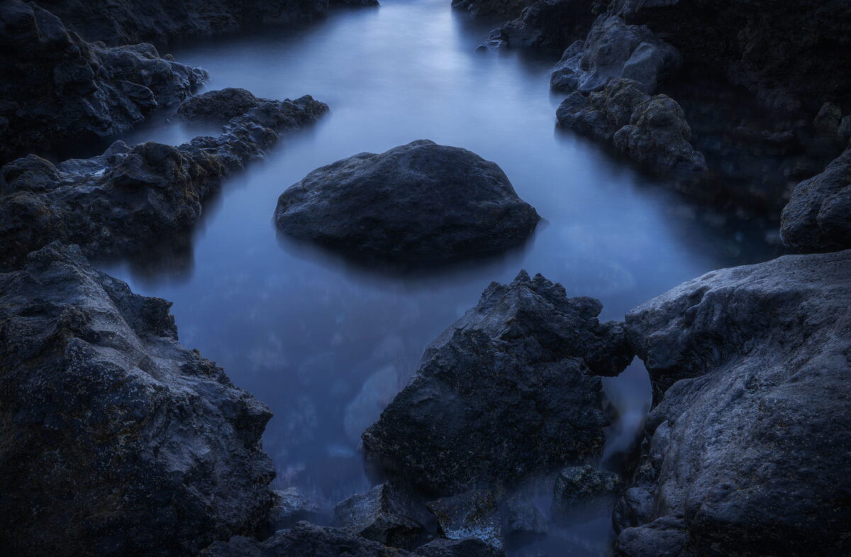 Long exposure seascape at Playa de Montaña Bermeja in Lanzarote, Canary Islands, with dark volcanic rocks surrounded by silky blue water during the blue hour.