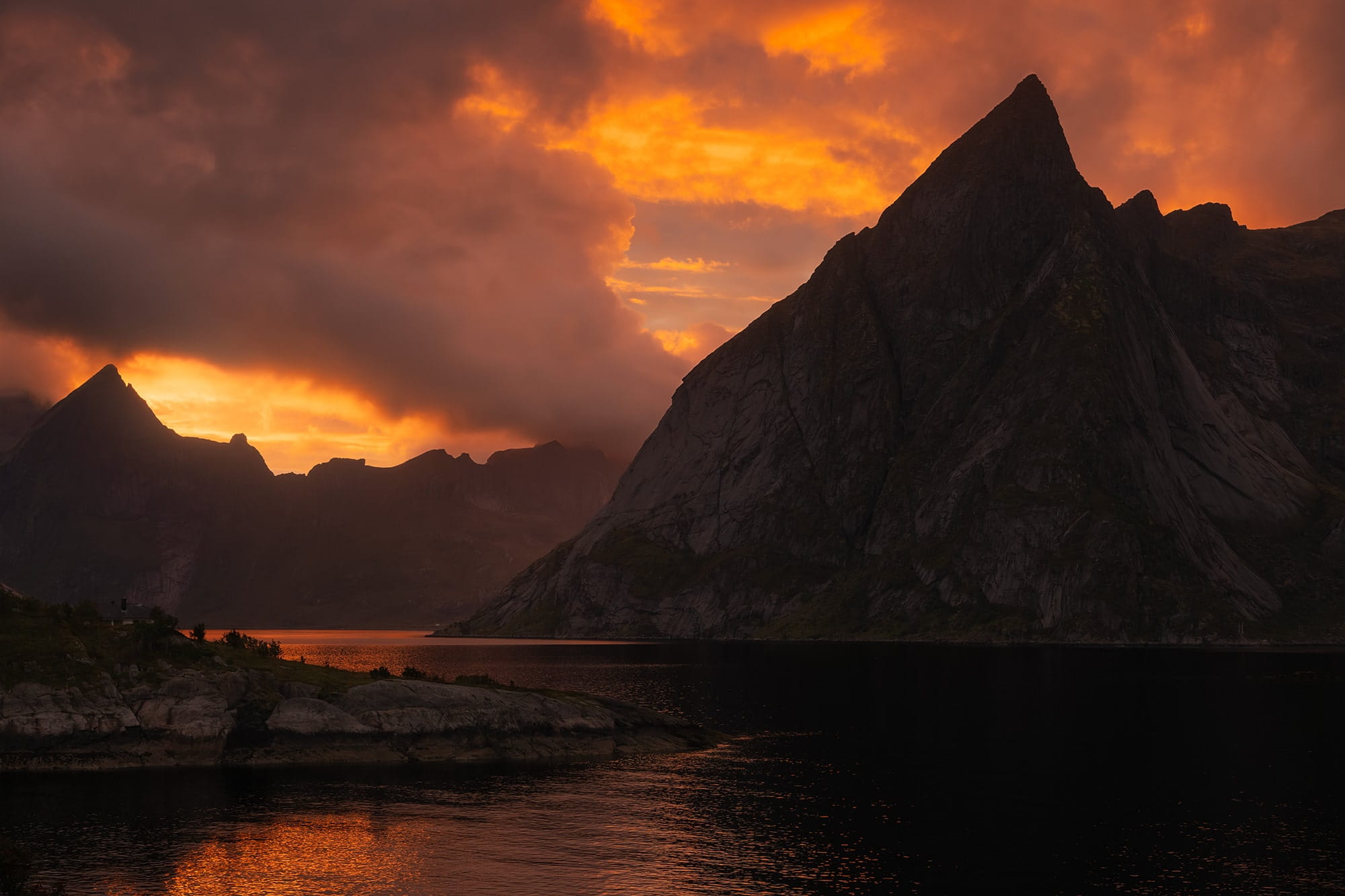 Dramatic sunset over Hamnøy in the Lofoten Islands, Norway, with jagged mountain peaks silhouetted against a fiery orange sky reflected in the fjord.