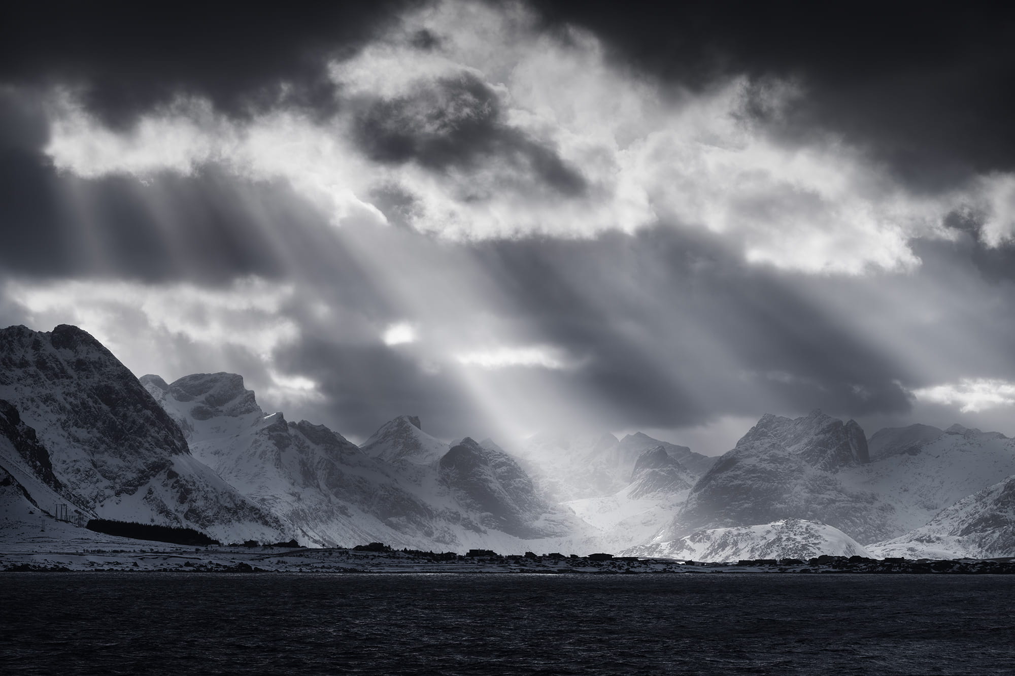 Dramatic sun rays breaking through storm clouds over snow-covered mountains in the Lofoten Islands, Norway, during winter.