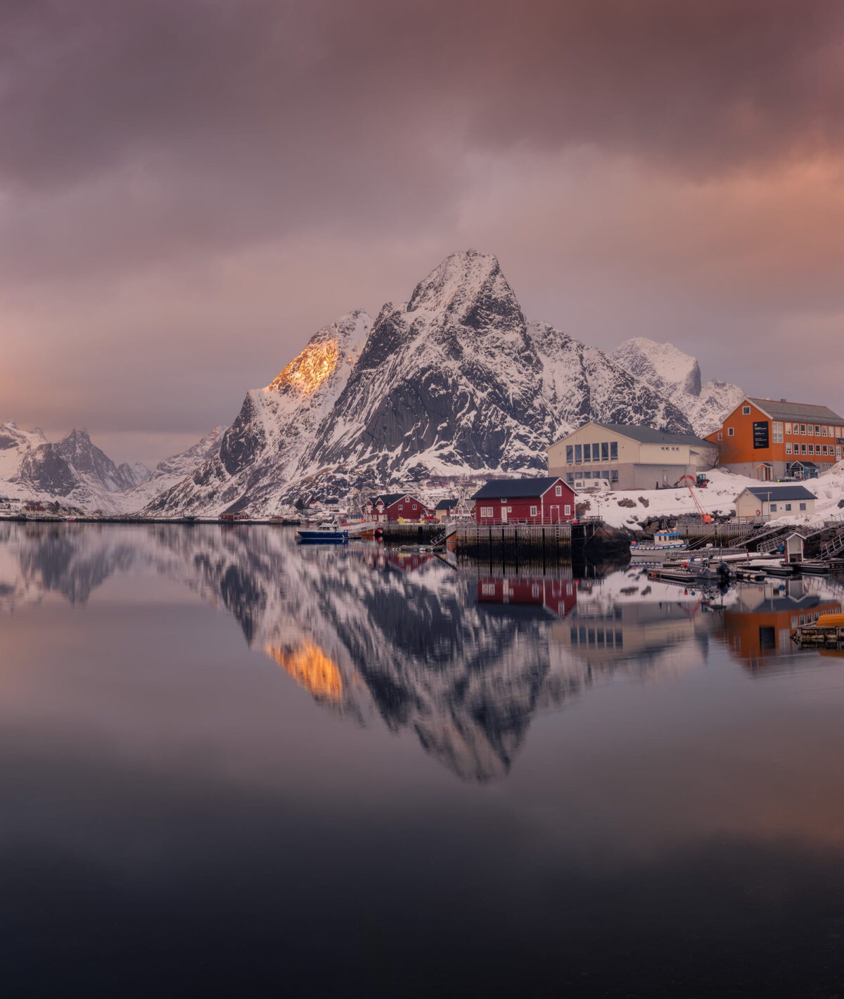 Snow-covered mountains and fishing cabins reflected in the calm waters of Reine, Lofoten Islands, Norway, during a soft winter sunset.