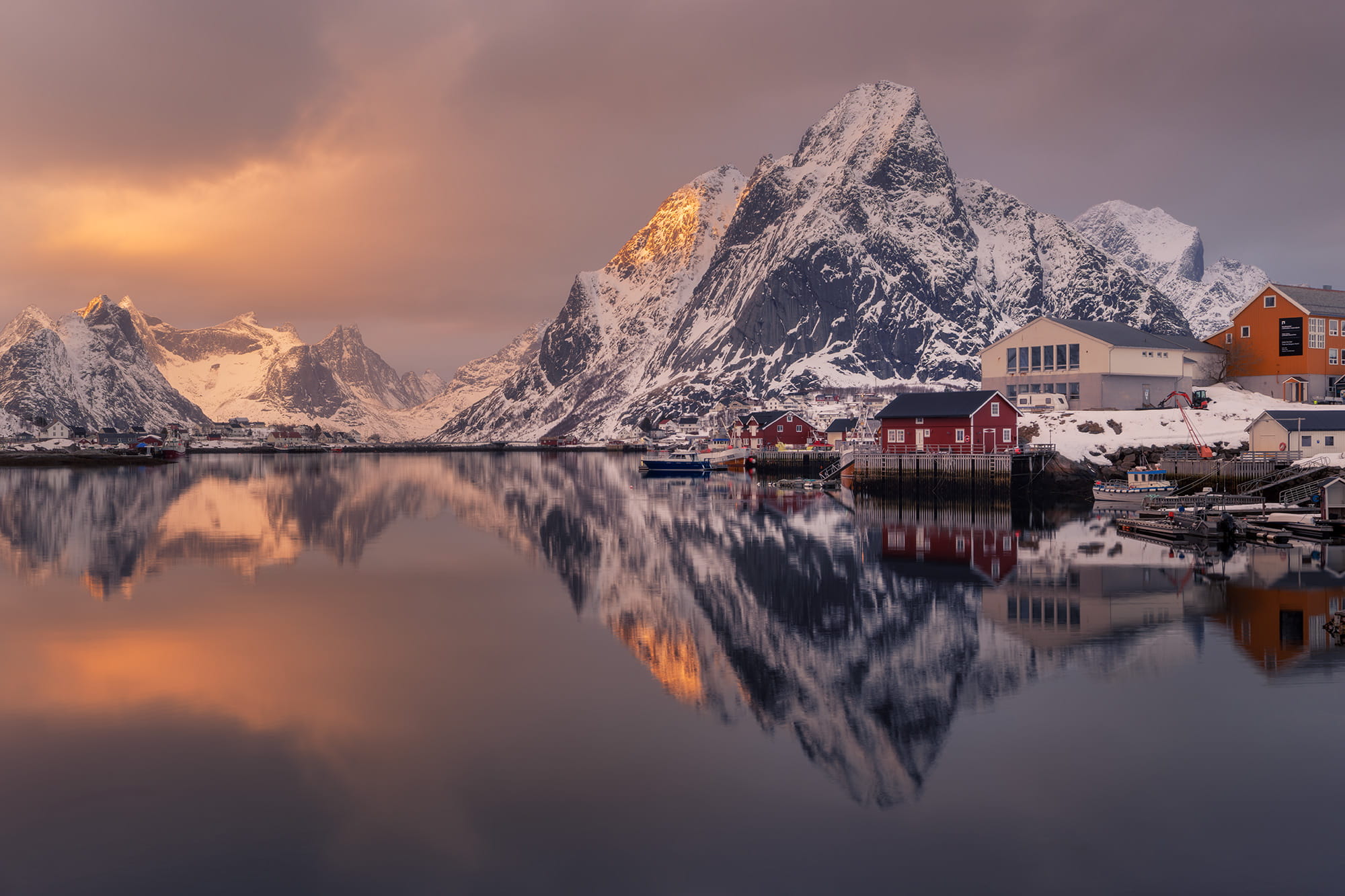 Winter landscape of Reine in the Lofoten Islands, Norway, with snow-covered mountains and traditional red fishing cabins reflected in the calm fjord at sunset.