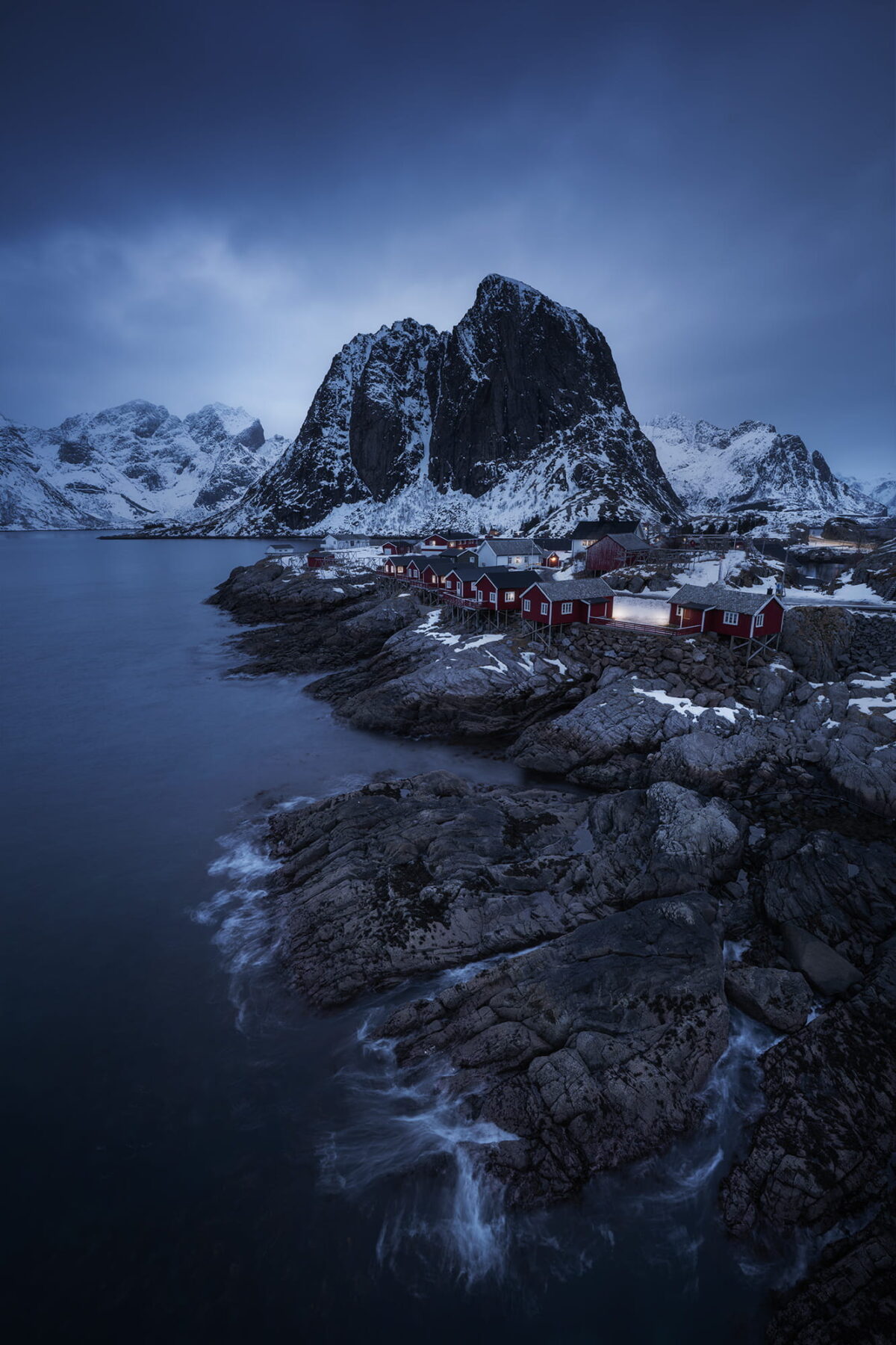 Winter landscape of Hamnøy in the Lofoten Islands, Norway, with traditional red rorbuer cabins along the rocky coast beneath dramatic snow-covered mountains at blue hour.