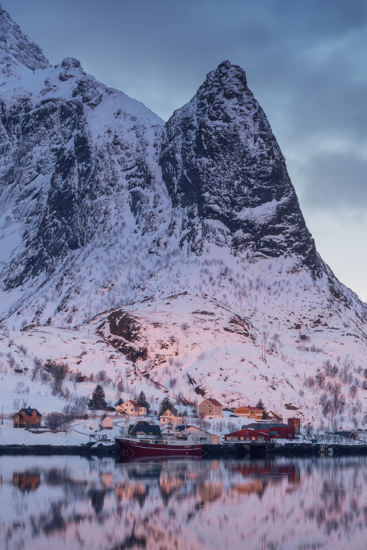 Fishing boat beneath a dramatic snow-covered mountain in Reine, Lofoten Islands, Norway, reflected in calm Arctic waters.