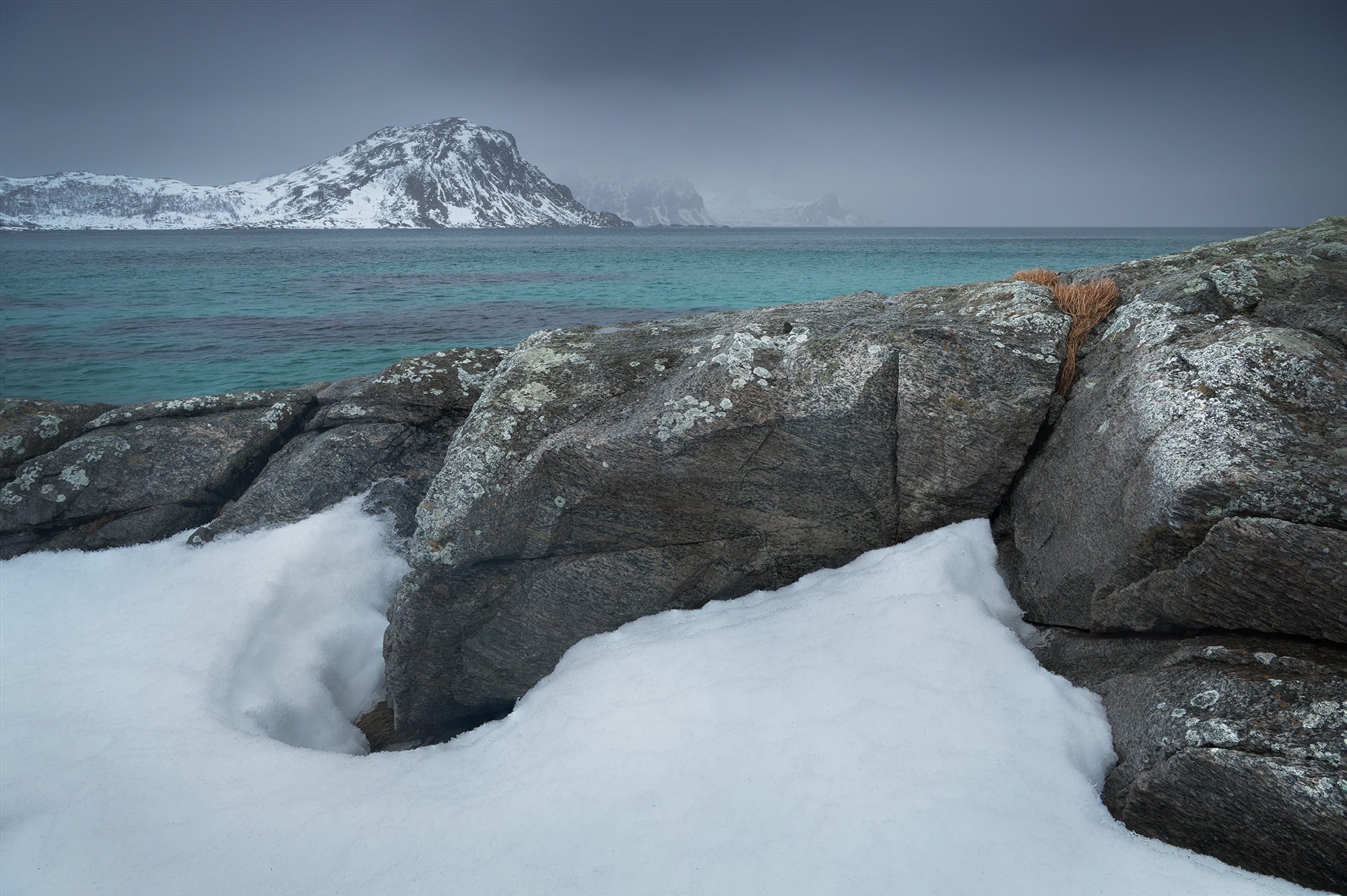 Snow-covered rocks and turquoise Arctic waters at Vik Beach in the Lofoten Islands, Norway, surrounded by snowy mountains in winter.