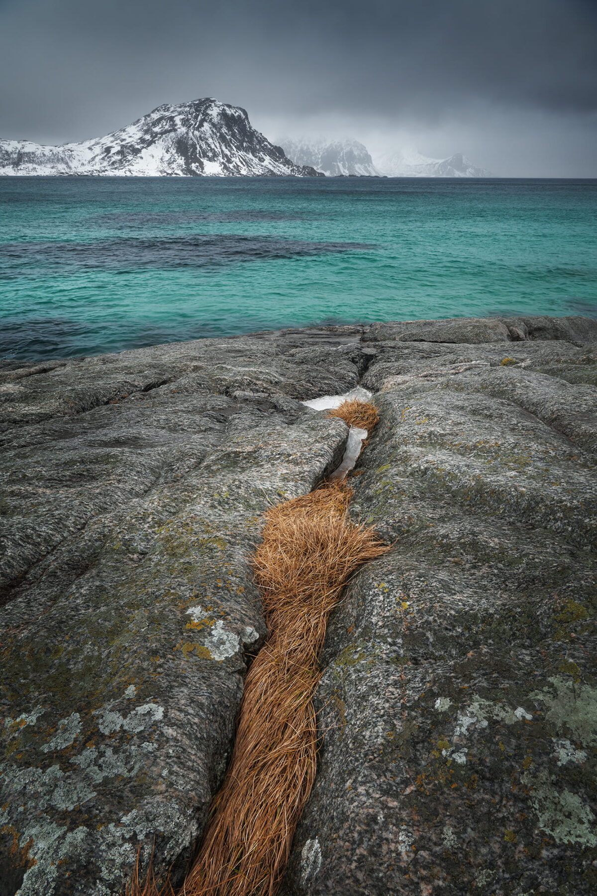 Winter landscape at Vik Beach, with turquoise Arctic water, rocky shoreline and snow-covered mountains in the Lofoten Islands, Norway.