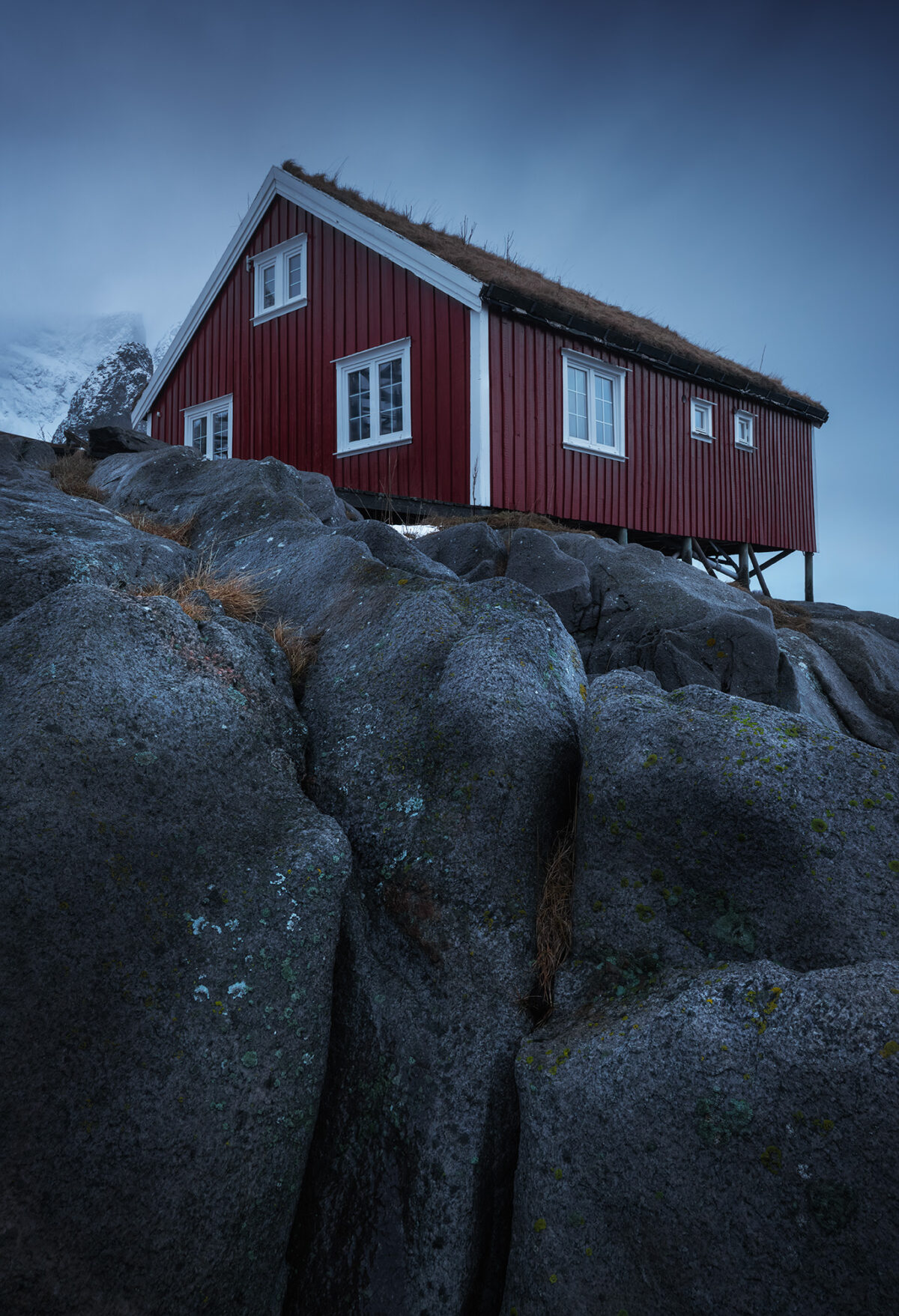 Traditional red rorbu cabin with a grass roof perched on rocky cliffs in Reine, Lofoten Islands, Norway, surrounded by a moody winter mountain landscape.
