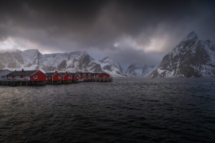 Red fishing cabins in Hamnøy, Lofoten Islands, Norway, surrounded by dramatic snow-covered mountains and dark Arctic waters in winter.