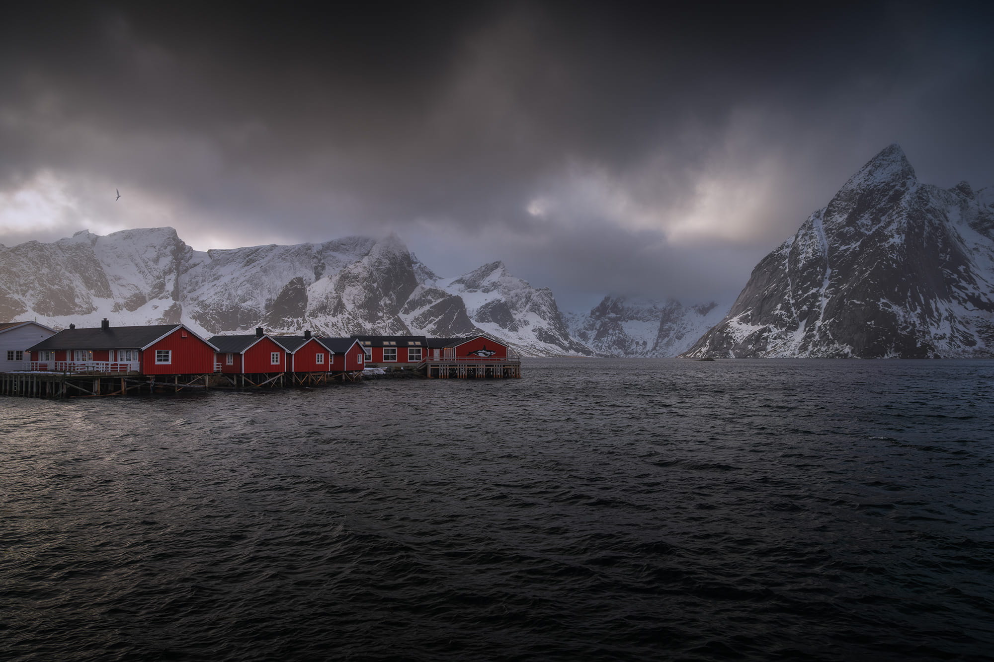 Red fishing cabins in Hamnøy, Lofoten Islands, Norway, surrounded by dramatic snow-covered mountains and dark Arctic waters in winter.