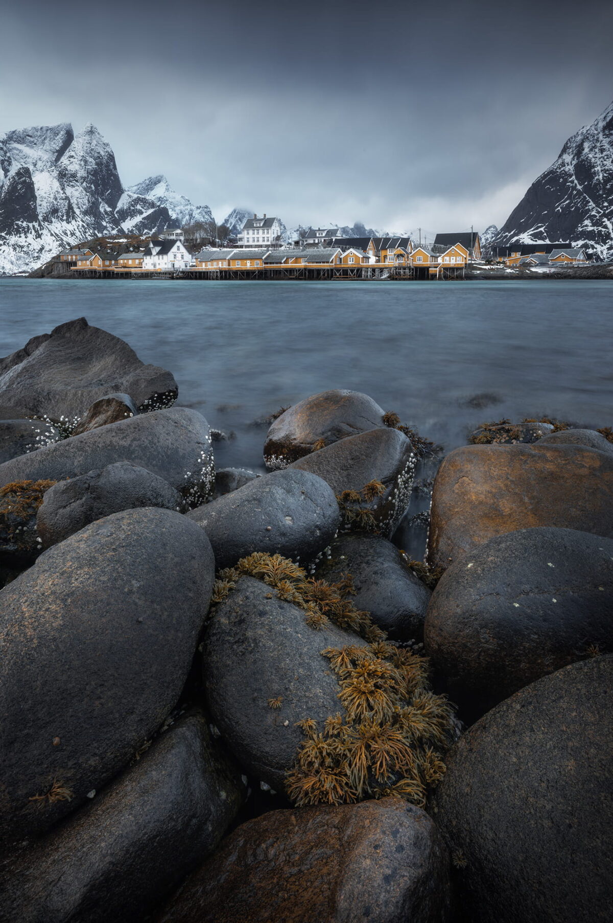 Traditional fishing village of Sakrisøy in the Lofoten Islands, Norway, surrounded by dramatic snow-covered mountains and viewed across the fjord from a rocky shoreline.