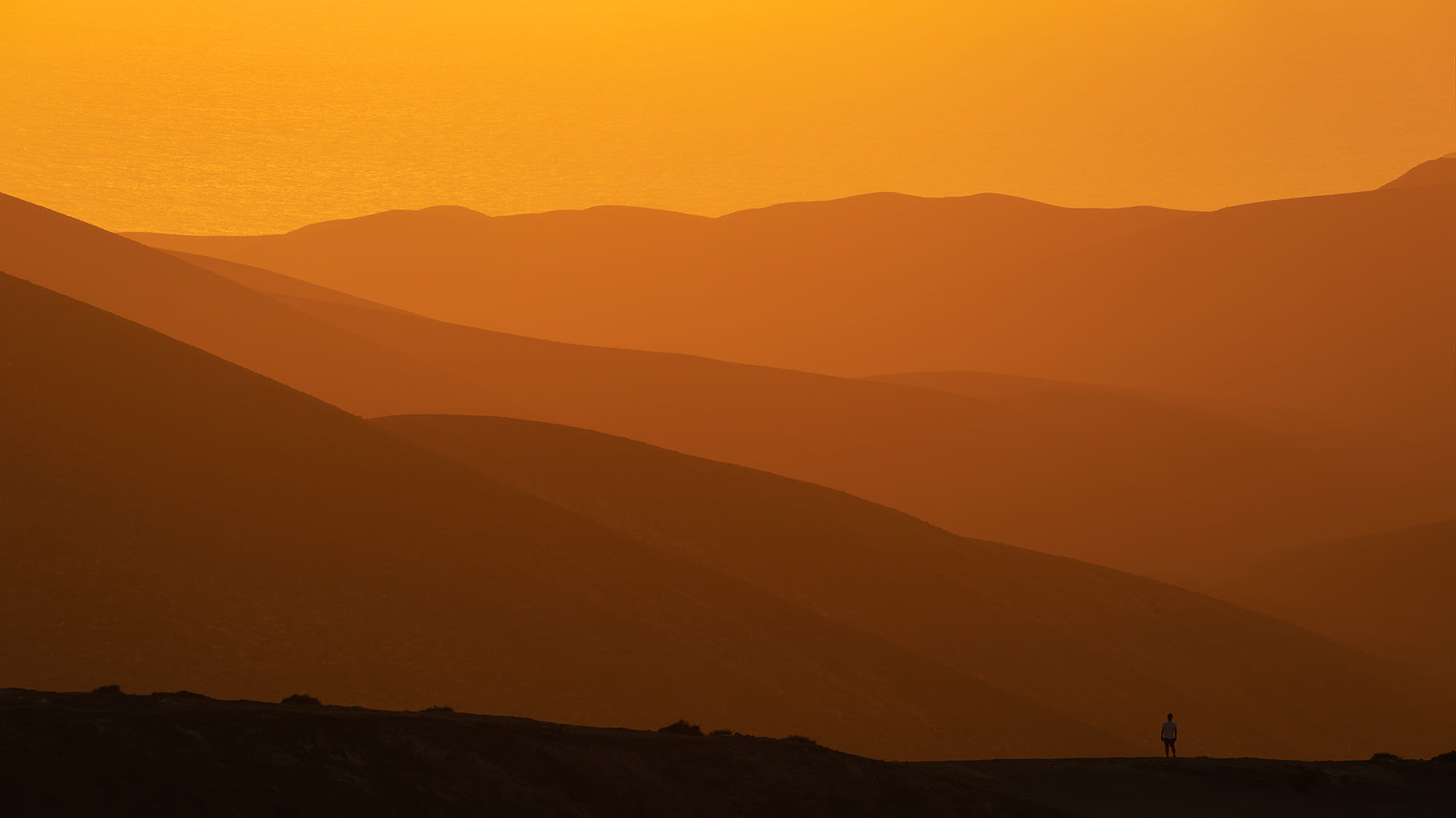 Layered volcanic mountains glowing at sunset from Mirador Astronómico de Sicasumbre in Fuerteventura, Canary Islands, with a lone person overlooking the landscape.