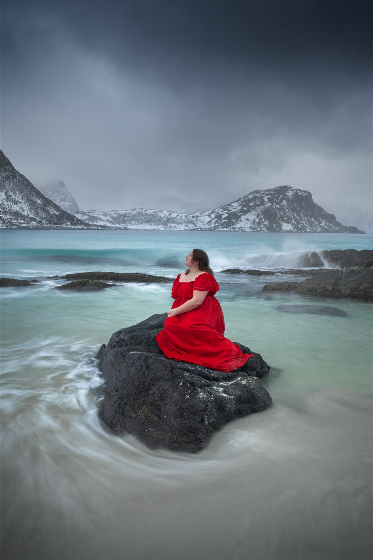 Woman in a flowing red dress on a rock at Vik Beach, surrounded by turquoise Arctic water and snowy mountains in winter in the Lofoten Islands, Norway.