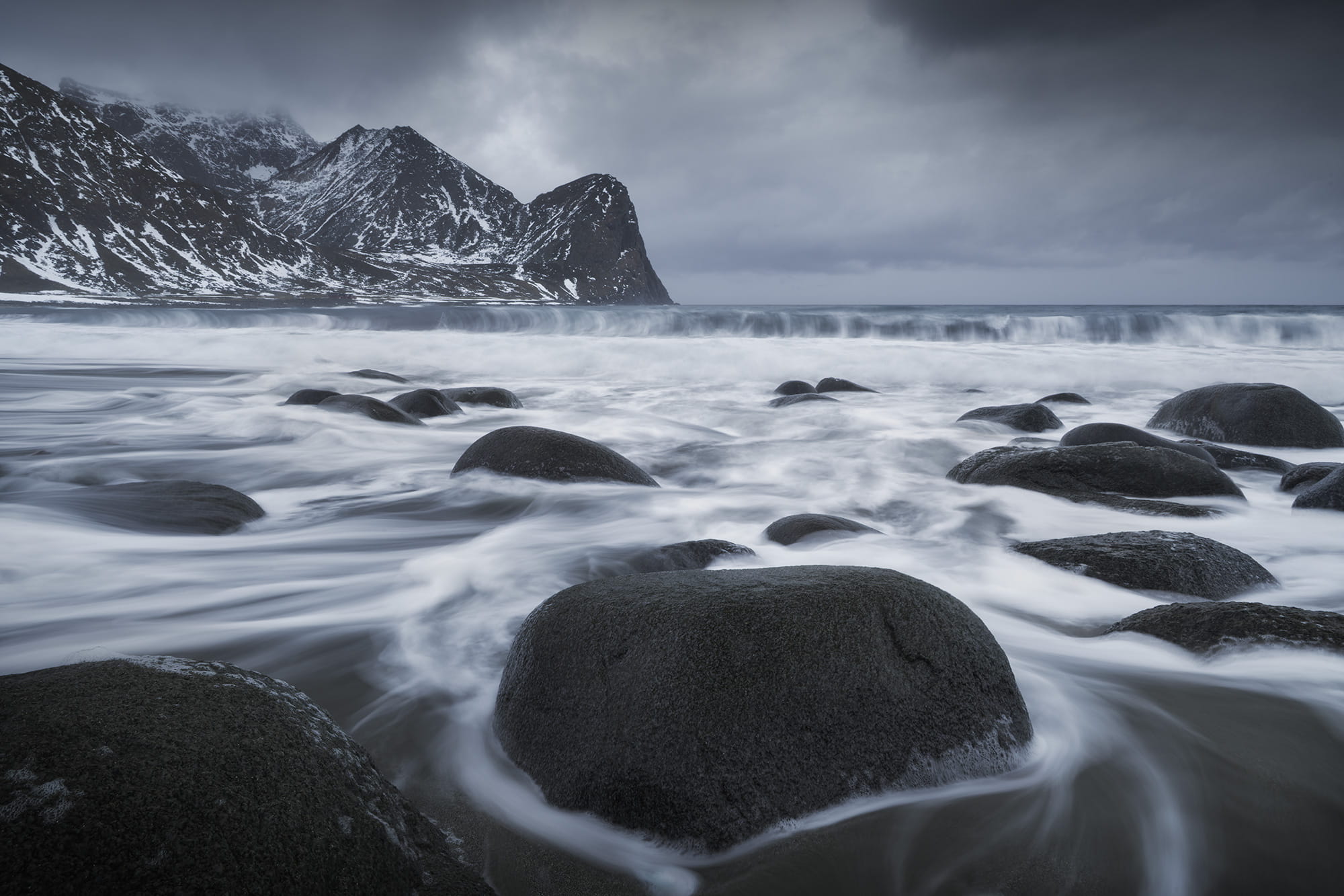 Long exposure of waves flowing around dark rocks at Unstad Beach in winter, beneath snow-covered mountains and a moody sky in the Lofoten Islands, Norway, photographed with a Nikon Z8.